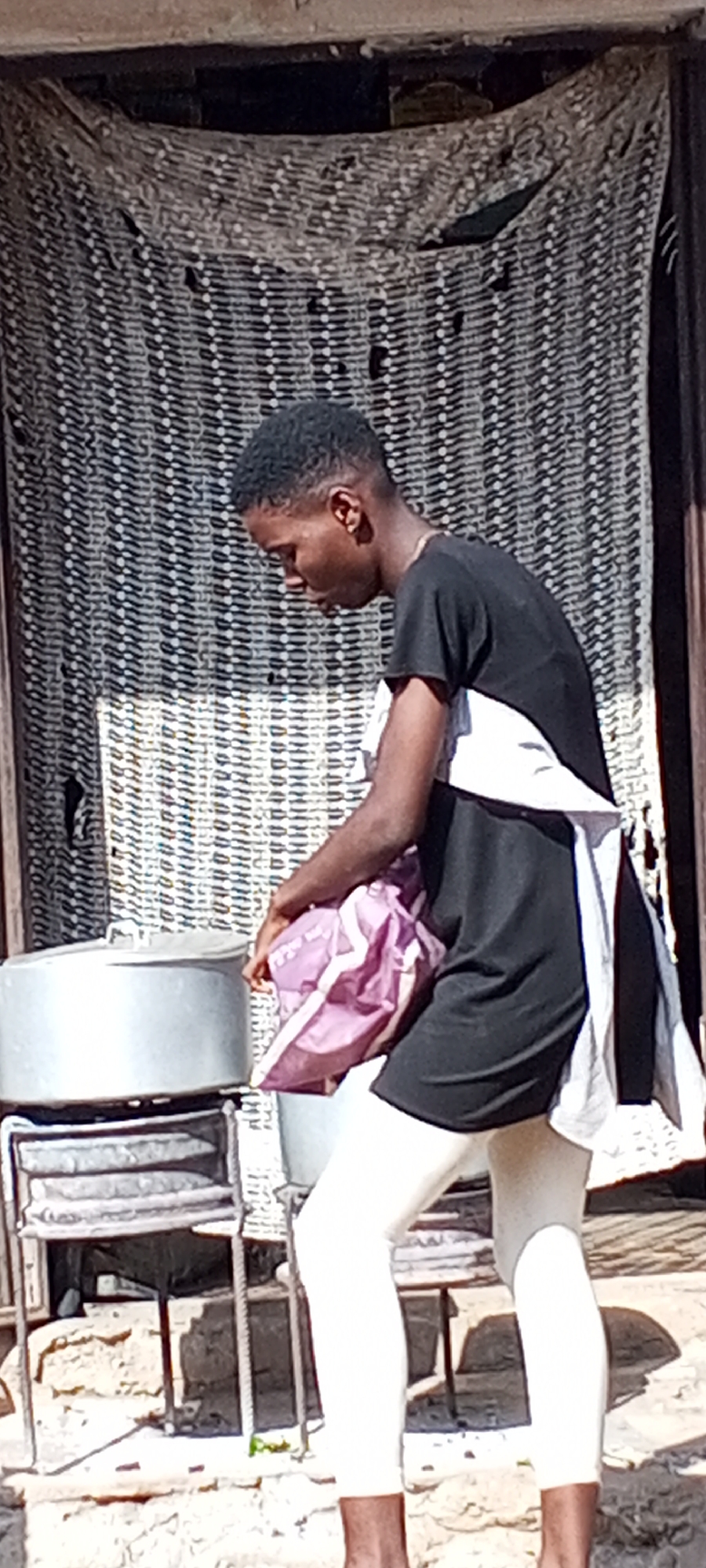 A young man stands in front of a building with a woven curtain in what appears to be an outdoor setting in Sabon Tasha, Nigeria. The man has short, dark hair and is wearing a black t-shirt, white leggings, and a white cloth draped over his shoulder. He is holding a pink shopping bag and looking down towards a large, silver pot on a metal stand. The building behind him has a rough exterior.