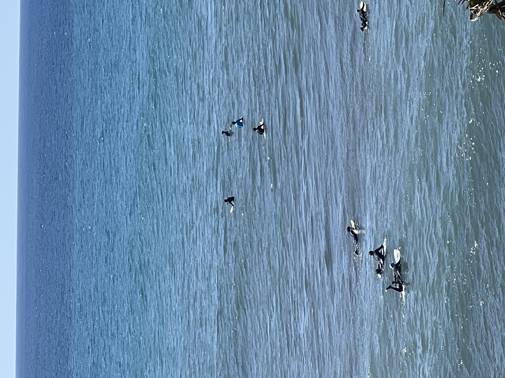 The image shows a section of the Atlantic Ocean off the coast of Maspalomas, Spain. Several surfers are visible scattered across the water, some paddling on their boards and others sitting and waiting for waves. The ocean surface appears textured with small waves and ripples. The weather seems clear and sunny, causing the water to glimmer. A portion of a wooden pier or structure is visible on the far right of the image. The sky is blue, and there are no boats or other watercraft in sight.