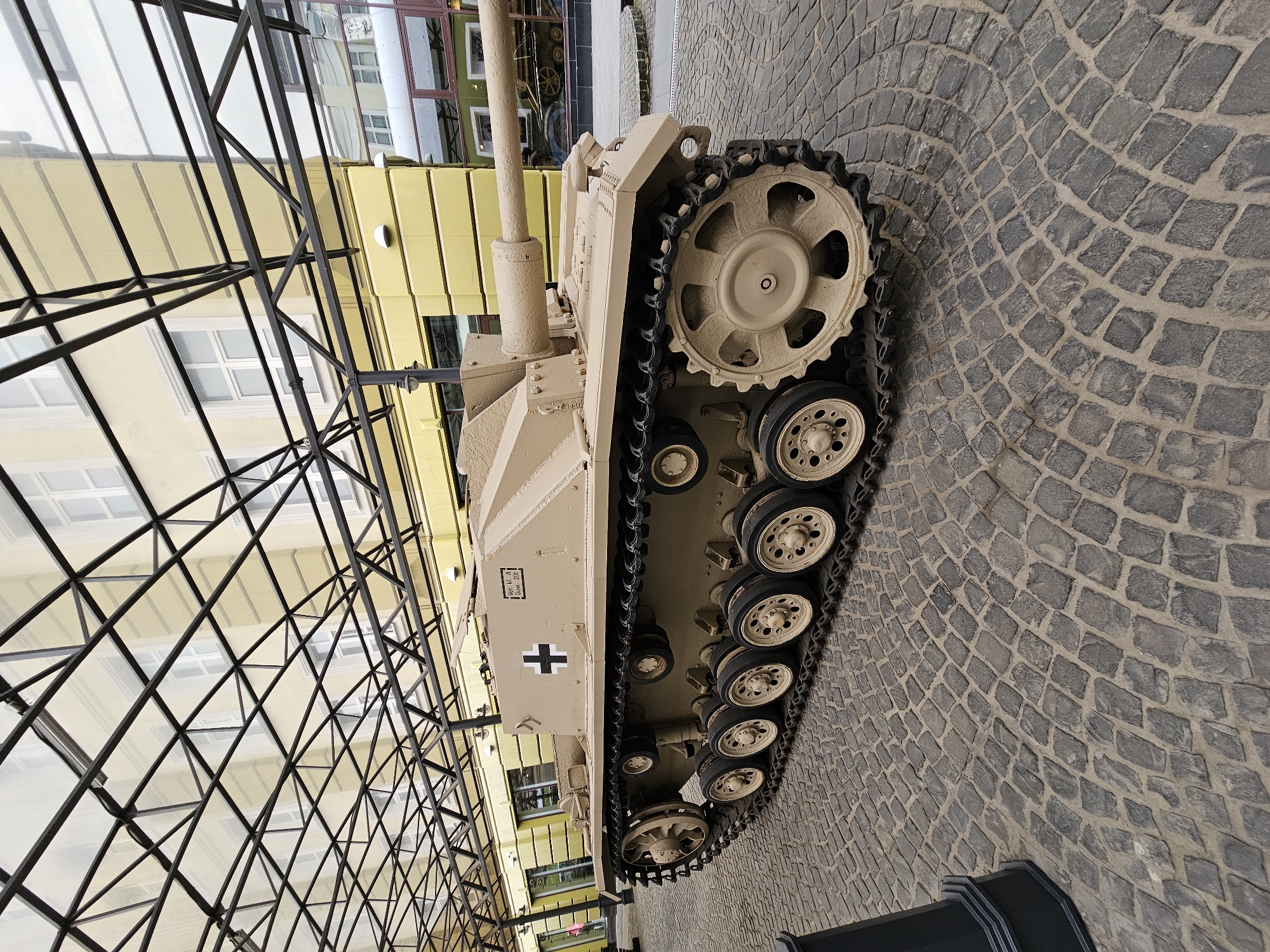 The image depicts a Panzerjäger 38(t) tank destroyer, likely a replica or restored vehicle, situated on a cobblestone surface. The tank is beige and features a black cross insignia. The scene appears to be an outdoor display, possibly within a museum or historical park. In the background, there are buildings reflecting in a glass ceiling with a metal frame. This aligns with the location Архангельское, Russia, which is known for its historical estate museum complex. The focus is on the tank itself, highlighting its features and placement within the environment.