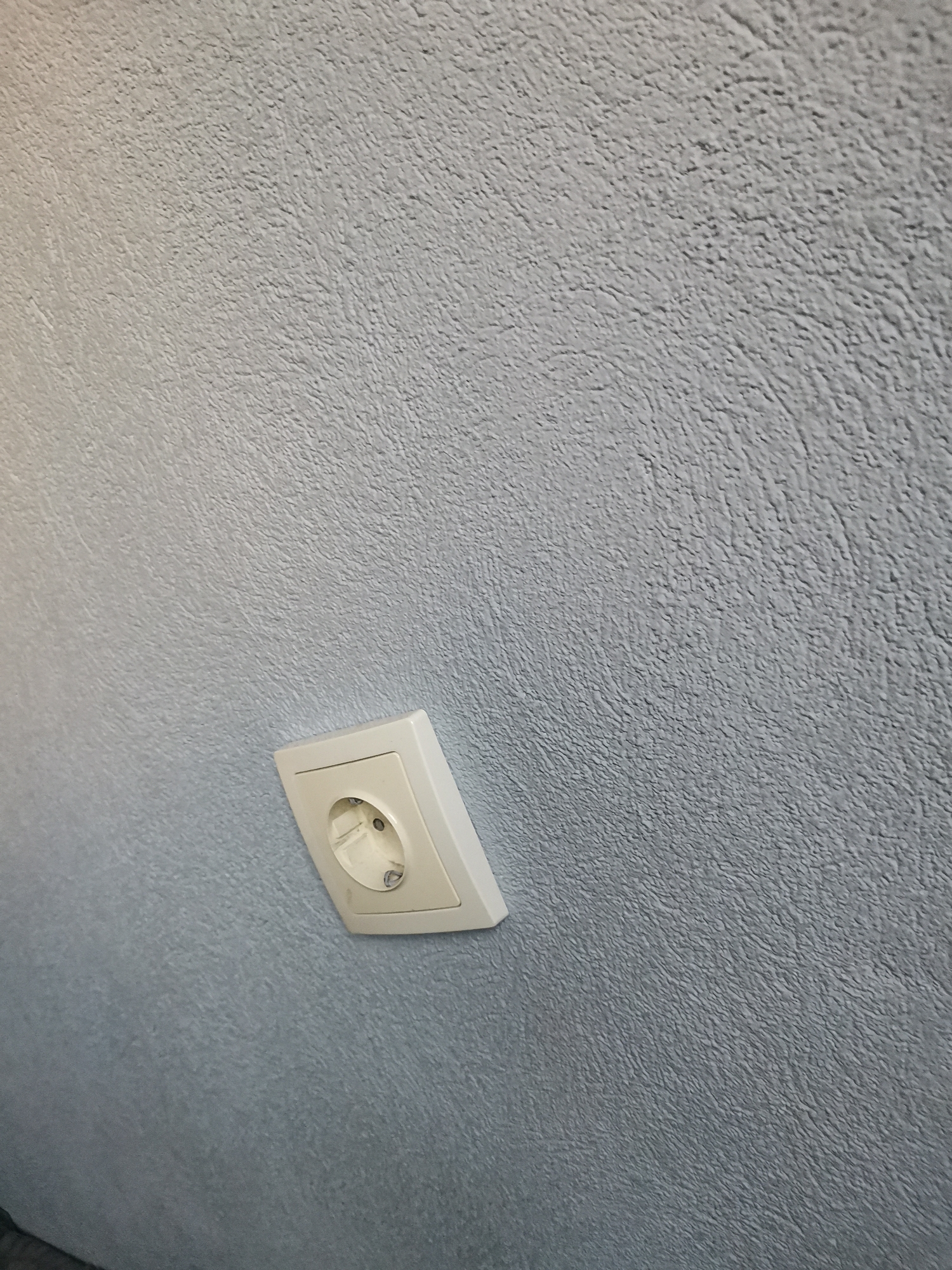 Located in Pokrovnik, Bulgaria, the image displays a textured wall rendered in a muted blue-grey tone. The wall surface features a coarse, irregular, and slightly undulating texture. A single beige or off-white electrical outlet, consistent with a Type F (Schuko) standard, is mounted on the wall. The outlet is oriented diagonally, with its top-right corner higher than its bottom-left. It presents two circular pin receptacles and a grounding clip, with minor dark marks visible inside the receptacles. Two screws secure the faceplate. The scene is static and devoid of human presence or activity.