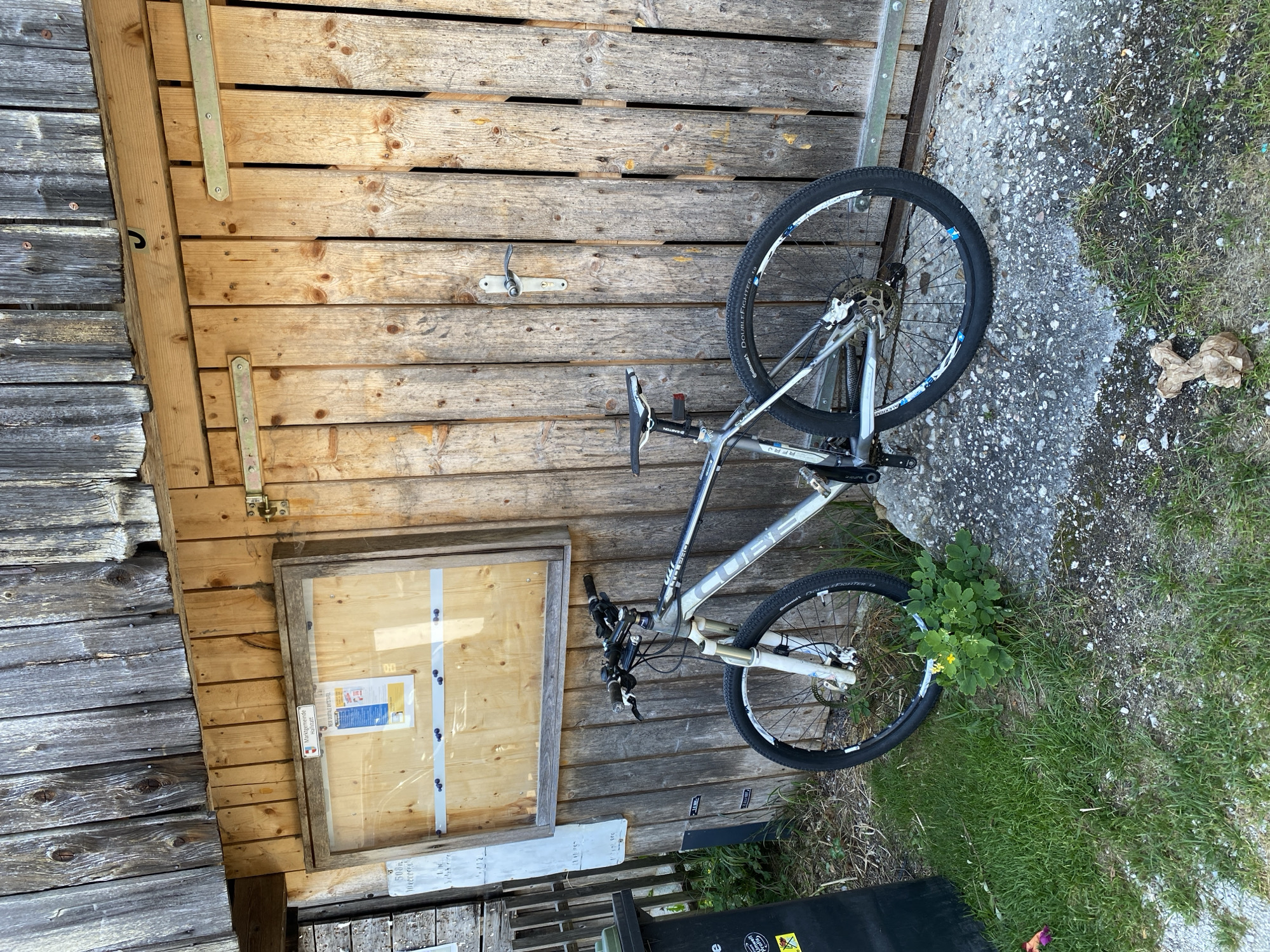 A silver and black mountain bicycle is parked against the weathered wooden wall of a building. The bicycle has a suspension fork and disc brakes. To the left of the bicycle is a black trash bin. A bulletin board with papers is mounted on the building wall above the bicycle. The ground in front of the building is a mix of grass and gravel. In the background, the wooden structure appears to be a shed or outbuilding. The scene is outdoors and appears to be in a rural or semi-rural setting. There are no people visible in the image. The lighting suggests it is daytime. Based on the "Marktgemeinde Hallstatt" sign visible on the bulletin board, this scene is located in the municipality of Hallstatt, Austria.