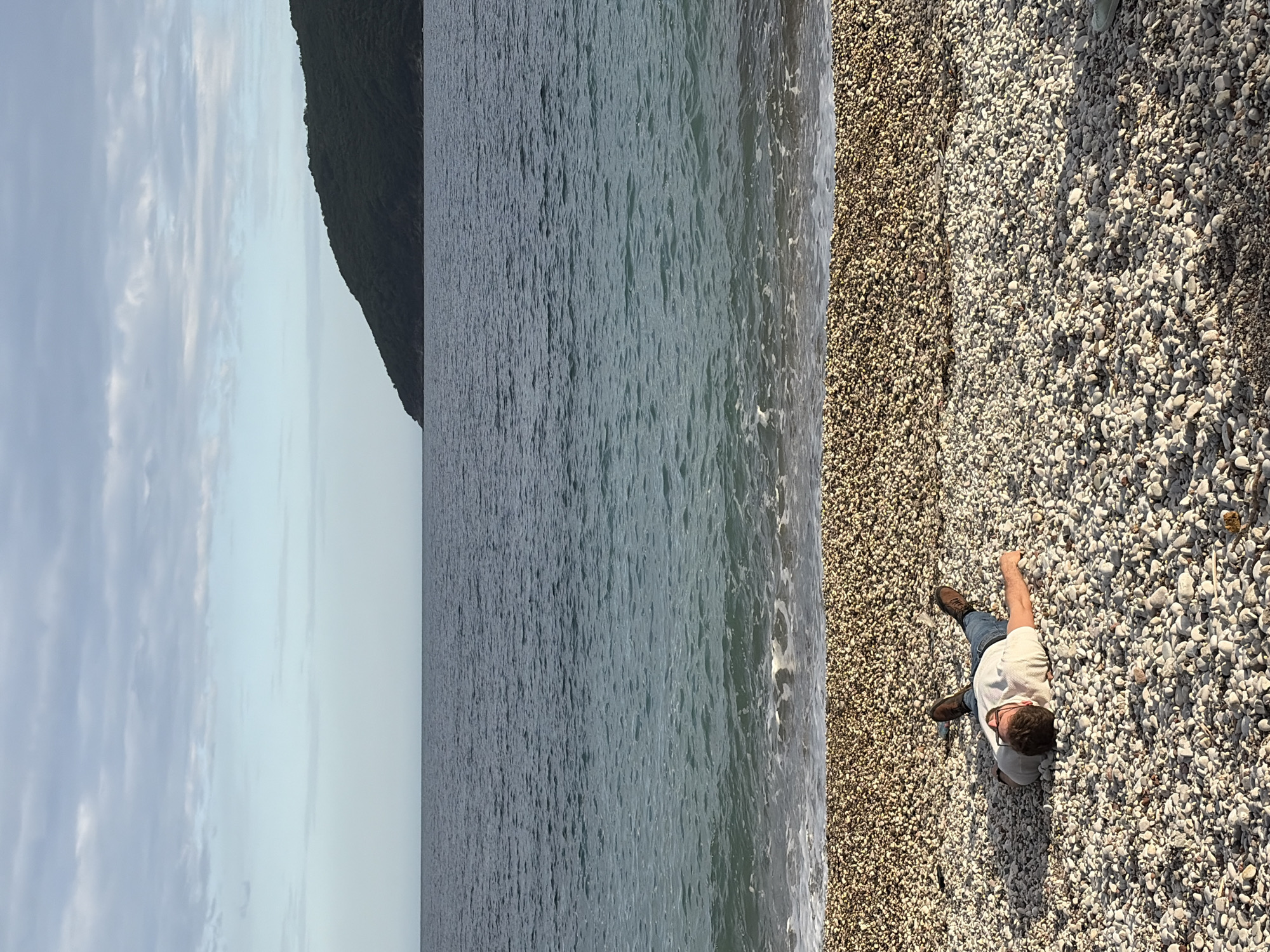A man is lying on a pebble beach next to the sea in Budva, Montenegro. The man is wearing a white t-shirt, blue jeans, and brown boots. He has dark hair and is wearing glasses. The sky is cloudy, and there is a mountain in the background. The water is blue-green.