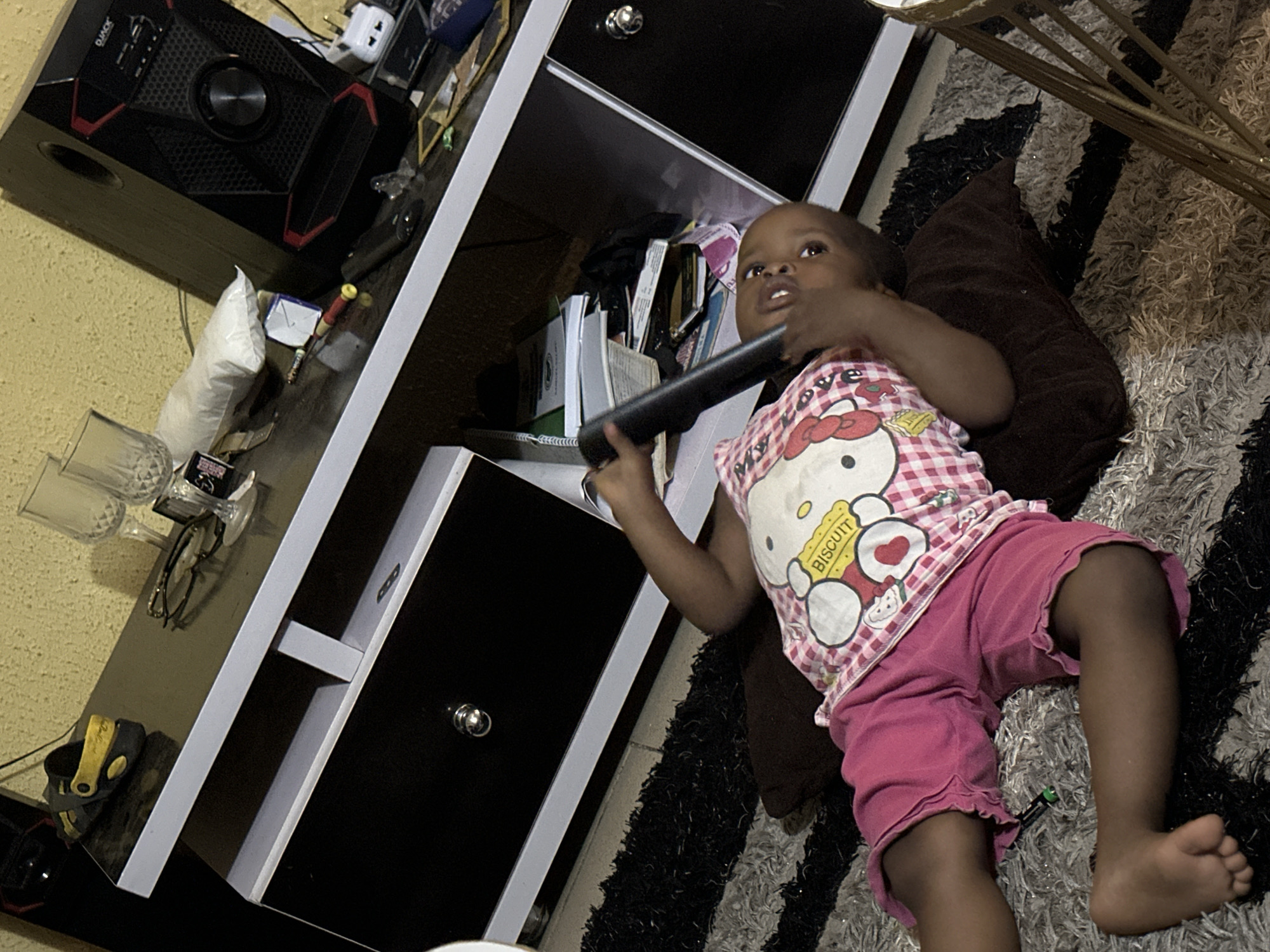 A young child, likely a girl, is lying on a patterned rug in what appears to be a living room in Narayi, Nigeria. The child is wearing a pink and white checkered shirt with a Hello Kitty graphic and pink shorts. She is holding a dark, cylindrical object, possibly a remote control or a toy, in her hands. Her gaze is directed upwards and to the right, suggesting she might be watching television or interacting with something off-camera.
Behind the child, a multi-tiered shelf unit with black drawers and white shelving is visible. The shelves are cluttered with various items, including books, papers, and what looks like a white plastic bag. To the left of the shelf unit, a black speaker system is positioned on a yellow surface, which may be a wall or another piece of furniture. On one of the shelves, a pair of glasses and some drinking glasses are placed. A dark-colored shoe or sandal is also visible on a lower surface.
The lighting in the scene suggests it could be daytime, possibly indoors with artificial lighting. The overall impression is a casual, home environment. There is no clear indication of the weather or specific time of day beyond the possibility of daylight. The child's expression is one of mild curiosity or engagement. No prominent text is clearly legible, apart from the "BISCUIT" label on the child's shirt.
