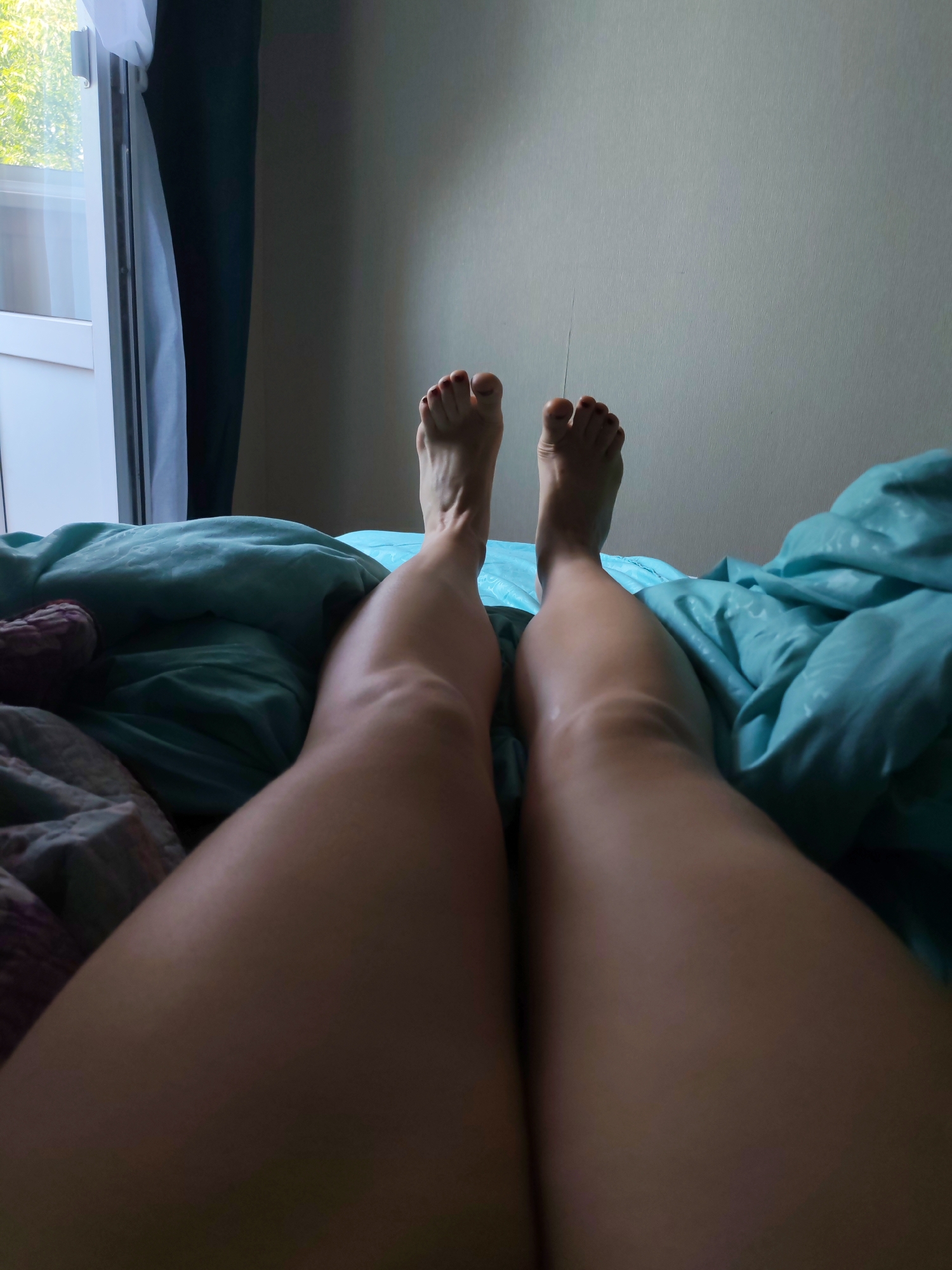 The image displays a pair of human legs, from the knees downwards, resting on a bed. The legs are bare and tanned, with toenails painted red. The bed is covered with a teal-colored comforter, and a patterned blanket is partially visible on the left. In the background, a window with sheer white and dark curtains is to the left, allowing natural light to illuminate the scene. The wall behind the bed is a neutral, light beige color. The perspective suggests the photo was taken from a low angle, looking up at the legs. The location is in Izhevsk, Russia, within a residential setting.