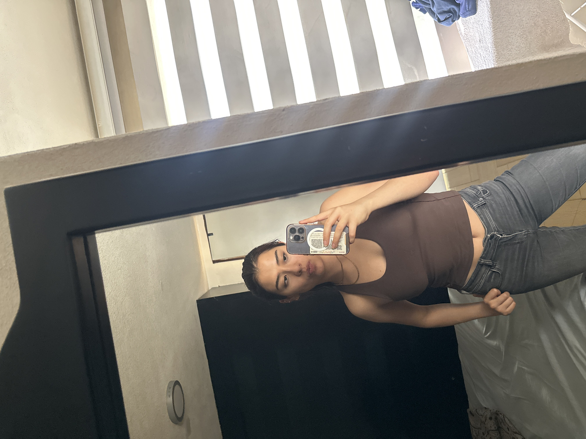 A young woman is captured in a mirror selfie, posing in what appears to be a bedroom or dressing area. The image is oriented sideways, with the woman's head tilted towards the left of the frame. She has dark hair styled back, a neutral expression with slightly pursed lips, and is holding a smartphone in her right hand to take the picture. She wears a fitted, sleeveless brown top and dark gray jeans with a visible silver button and zipper. A thin gold-colored chain necklace is also visible around her neck.
The mirror she is using has a thick black frame. Reflected in the mirror, the background reveals light-colored walls, possibly off-white or beige. In the upper background, vertical window blinds or slats are partially open, allowing bright light to stream in and creating a striped pattern of light and shadow, indicating it is daytime. On the right side of the reflection, part of a bed with white bedding or a comforter is visible, along with a patterned blanket or throw underneath. A dark piece of furniture, possibly a dresser or cabinet, is reflected behind her on the left, and a small, circular, silver-rimmed object, resembling a light switch or thermostat, is mounted on the wall. A blue piece of cloth is loosely visible in the top right corner of the reflection. The smartphone she holds has multiple camera lenses and white stickers or notes adhered to its back, with some illegible text and barcode-like patterns. The scene suggests an indoor setting in Puebla, Mexico.