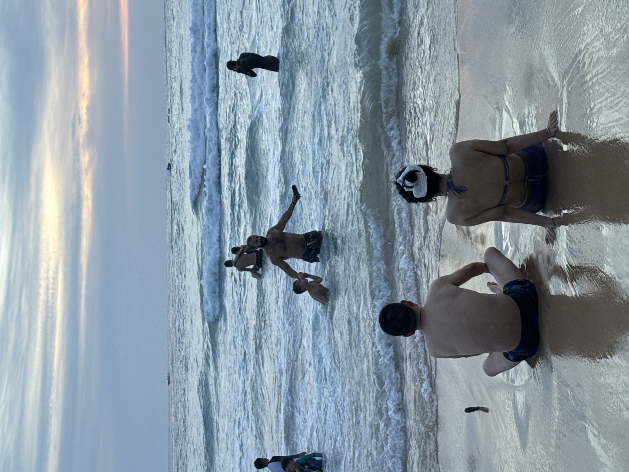 The image depicts a beach scene with several people in and around the water at Karon, Thailand. The sky is overcast with streaks of pale orange and pink, suggesting either sunrise or sunset. The sea is relatively calm with gentle waves rolling onto the shore.
In the water, there are multiple individuals. Two people are near the foreground, sitting on the wet sand where the water meets the beach. One, viewed from behind, is wearing dark swim shorts. The other, also viewed from behind, is wearing a bikini and has a white item on their head. Further out in the water, a man is partially submerged, with his arm extended towards a child who is also in the water. Another person is visible in the water further back, appearing to be lying down or floating. There's also a person in black swimwear floating face down in the distance.
The overall mood is relaxed, with people enjoying the water and the beach. The lighting is soft, consistent with either early morning or late evening. The wet sand reflects the light and the sky, creating a glossy texture. There is no visible text in the image.