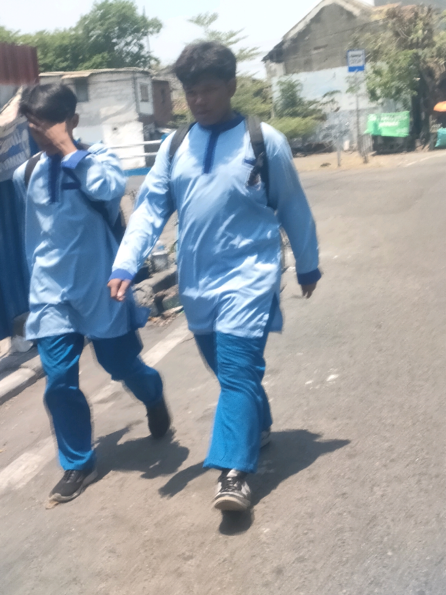 In Surabaya, Indonesia, two young males in blue uniforms walk on a paved street. The male in the foreground, with dark hair, wears a light blue tunic with a dark blue collar and matching blue pants. He has a black backpack slung over his left shoulder and black and white sneakers. The other male, partially obscured by the first, also wears a similar blue uniform and backpack and is walking to the left. Behind them, a white building with a corrugated metal roof is visible, as are trees and a street sign. The ground is asphalt, and shadows are cast on the road. The scene appears to be a sunny day.