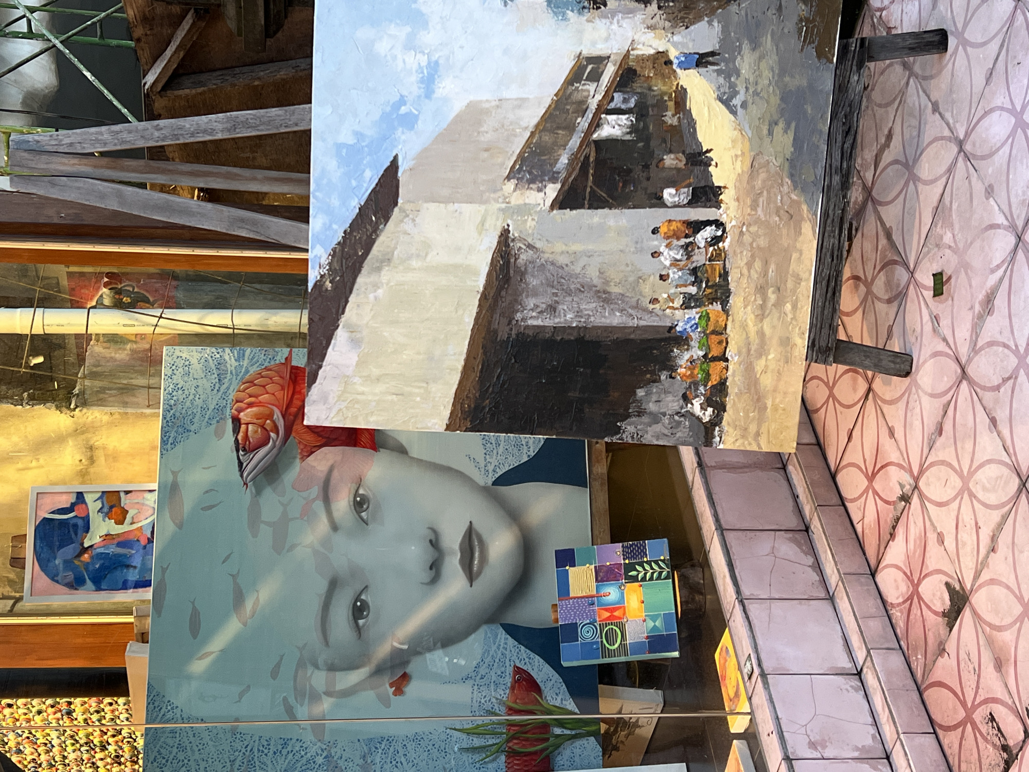 This media file showcases an outdoor or semi-outdoor display of two artworks, likely within a gallery or shop setting in Ubud District, Indonesia.
The upper painting, rendered with visible brushstrokes, depicts a vibrant street or market scene. A large, light-colored building with dark rooflines and windows dominates the background under a light blue sky with wispy clouds. Numerous small, stylized figures, some appearing to be people in traditional attire engaged in trade or daily life, are visible on a dirt path in the foreground. This painting rests on a rough wooden easel or support.
Below it, a more detailed painting features the serene face of a woman with subtle Asian features. Her head is adorned with an elaborate, textured red element resembling coral or intricate hair, and small fish motifs appear to swim around her face against a light blue, patterned background. Reflections on the protective glass of this painting reveal other elements: a smaller framed abstract artwork with blue and pink shapes to the left, and a partial view of a display of numerous colorful, spherical objects (possibly beads or decorative items) at the bottom left. A small, colorful, patchwork-like decorative object is also visible on the right side of the reflection.
To the right, a pinkish-tiled floor with a repeating circular geometric pattern is prominent. Weathered wooden structures and beams are visible at the top and extending from the left, suggesting a rustic or traditional architectural context. The scene is brightly lit, indicative of daytime. No visible text is discernible in the image.