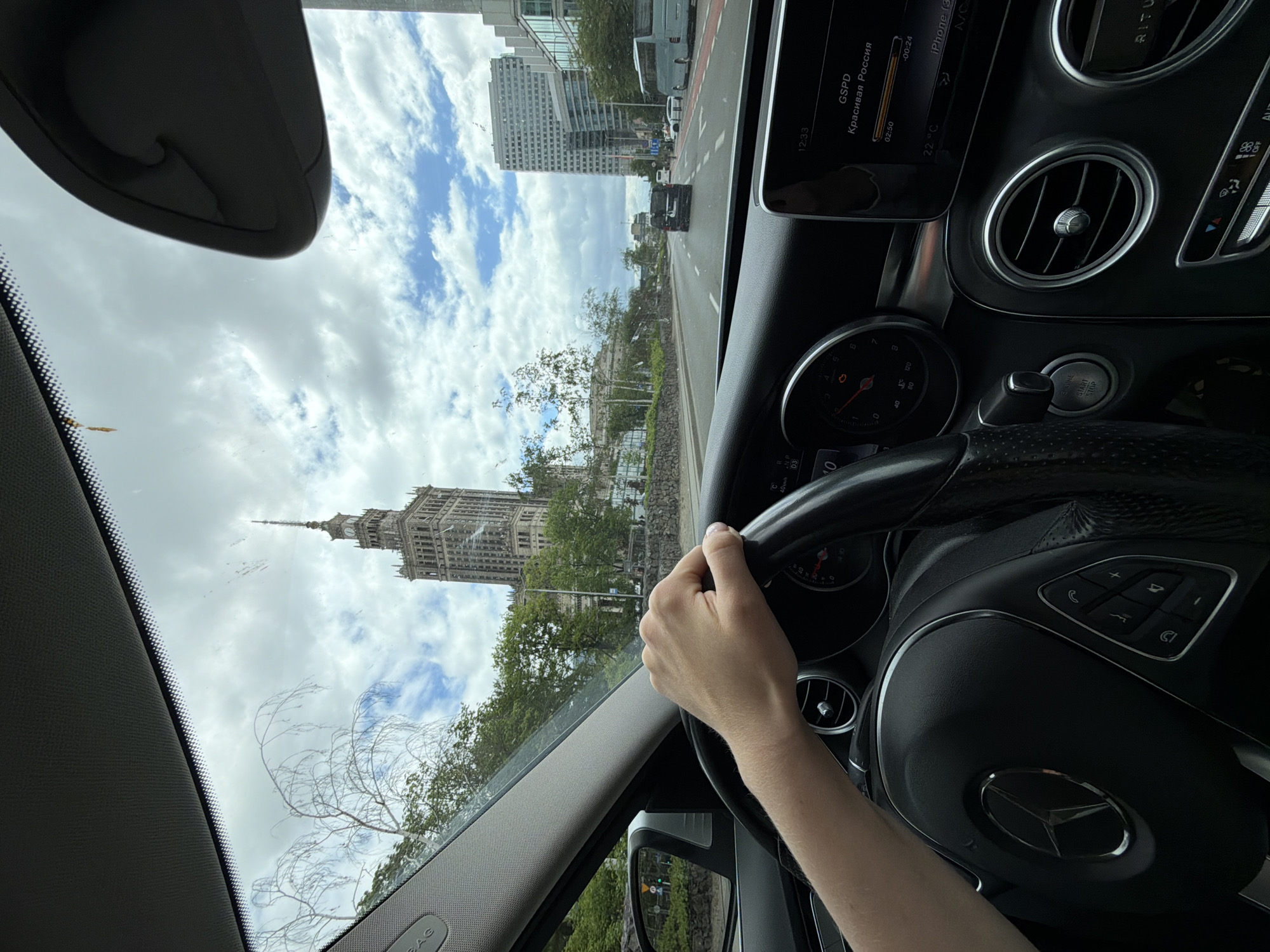 The image shows a view from inside a Mercedes-Benz car, likely a C-Class model, as the driver navigates a city street. The driver's left hand is visible on the steering wheel, and the dashboard and instrument panel of the car are in focus. The car's infotainment screen displays information in Russian, including the time and temperature. Through the windshield, there is a view of a city skyline with a tall, ornate building with a spire, which is the Palace of Culture and Science in Warsaw, Poland. Other modern buildings and trees line the street, and the sky is partly cloudy. Other vehicles can be seen on the road ahead.