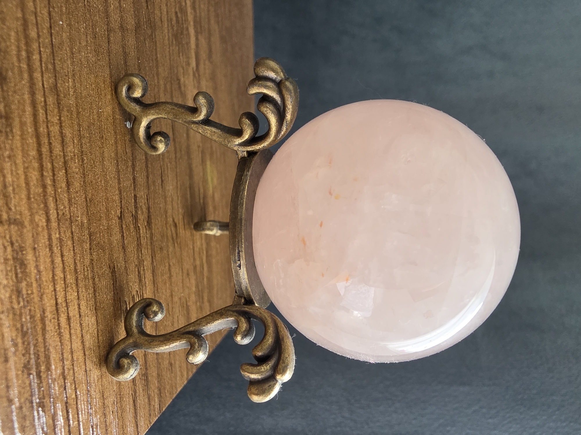A pink crystal sphere rests on an ornate brass stand, which is placed on a wooden surface. The background is a dark, textured material. The lighting is soft, highlighting the sphere's transparency and internal inclusions. The scene appears to be indoors, possibly a home or a shop, with no specific identifiable landmarks of Poprad, Slovakia.