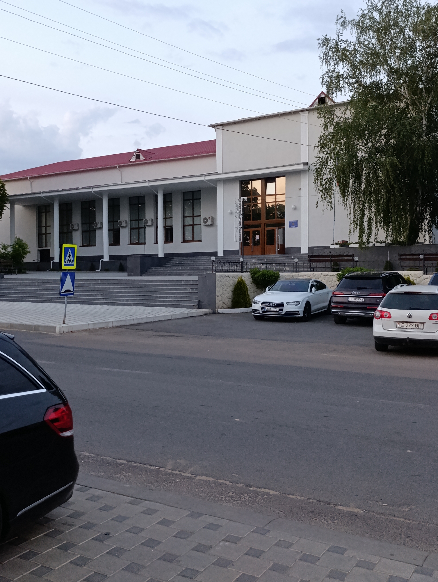 The image shows a building with a red roof and a row of columns along the front. A set of stairs leads up to the entrance. In front of the building, there are several cars parked on a paved area. A pedestrian crossing sign and a speed bump sign are visible near the stairs. A black car is partially visible on the left side of the image, parked on the asphalt road. The sky is cloudy. The building is located in the city of Cocieri, Moldova.
