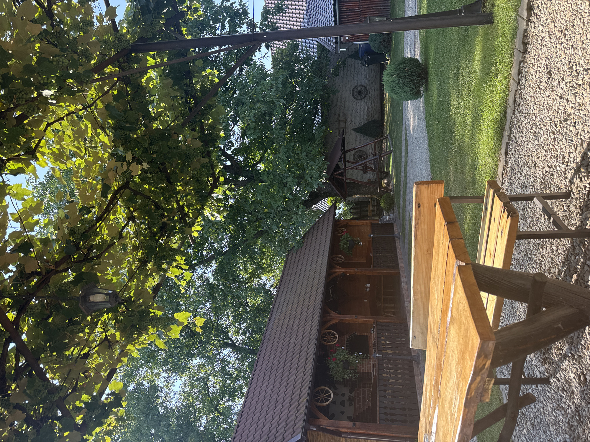 The image depicts an outdoor seating area at a rustic establishment in Cârțișoara, Romania. A wooden picnic table with benches occupies the foreground, positioned on a gravel surface. Behind the table, a wooden porch with a tiled roof extends from a building. This building appears to be a covered pavilion with wooden columns and decorative wagon wheels. In the background, a large tree with abundant green leaves shades a grassy area. A wooden swing set and a brick building with a tiled roof are also visible. A person is partially visible near the brick building, and a trash bin is located next to it. The scene is bathed in natural sunlight, suggesting daytime. The overall impression is one of a tranquil, rural setting, possibly a guesthouse or restaurant.