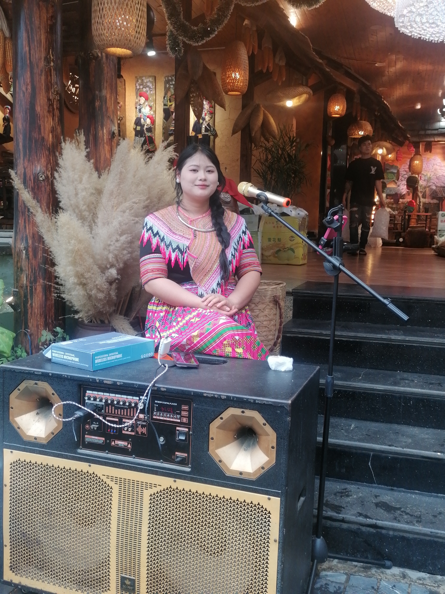 This full-shot image captures a young woman, likely of an ethnic minority group in Sa Pa Ward, Vietnam, sitting in what appears to be a semi-outdoor or covered shop entrance, poised for a performance. She is adorned in a vibrant, intricately patterned traditional outfit featuring geometric designs in black, pink, red, and white, with a silver-colored neck ring. Her dark hair is styled in a single braid, and she looks directly at the camera with a calm, pleasant smile. She is seated on a small woven stool, flanked by a large arrangement of dried ornamental pampas grass.
In front of her, a large black speaker system is set up, featuring gold-colored horn speakers and a gridded speaker area at the bottom, labeled "QC-PRO." The control panel of the speaker is visible, displaying "SENIOR DIGITAL PLAYER" and "BLUE," along with smaller labels "Y" and "B". A gold-tipped microphone on a black stand is positioned centrally. On top of the speaker, a blue box labeled "PROFESSIONAL UNIVERSAL WIRELESS MICROPHONE" is placed, alongside a plugged-in smartphone and a small white item.
The background features rustic wooden pillars and overhead structures adorned with woven lanterns and hanging corn cobs. Decorative wall hangings depicting figures in traditional attire are also visible. In the deeper background, another man in a black t-shirt stands near steps. A yellow box with the text "雪花梨" (Snow Pear) is partially visible. The scene, with its traditional elements and performance setup, suggests a cultural showcase or entertainment in a local market or tourist area in Sa Pa.