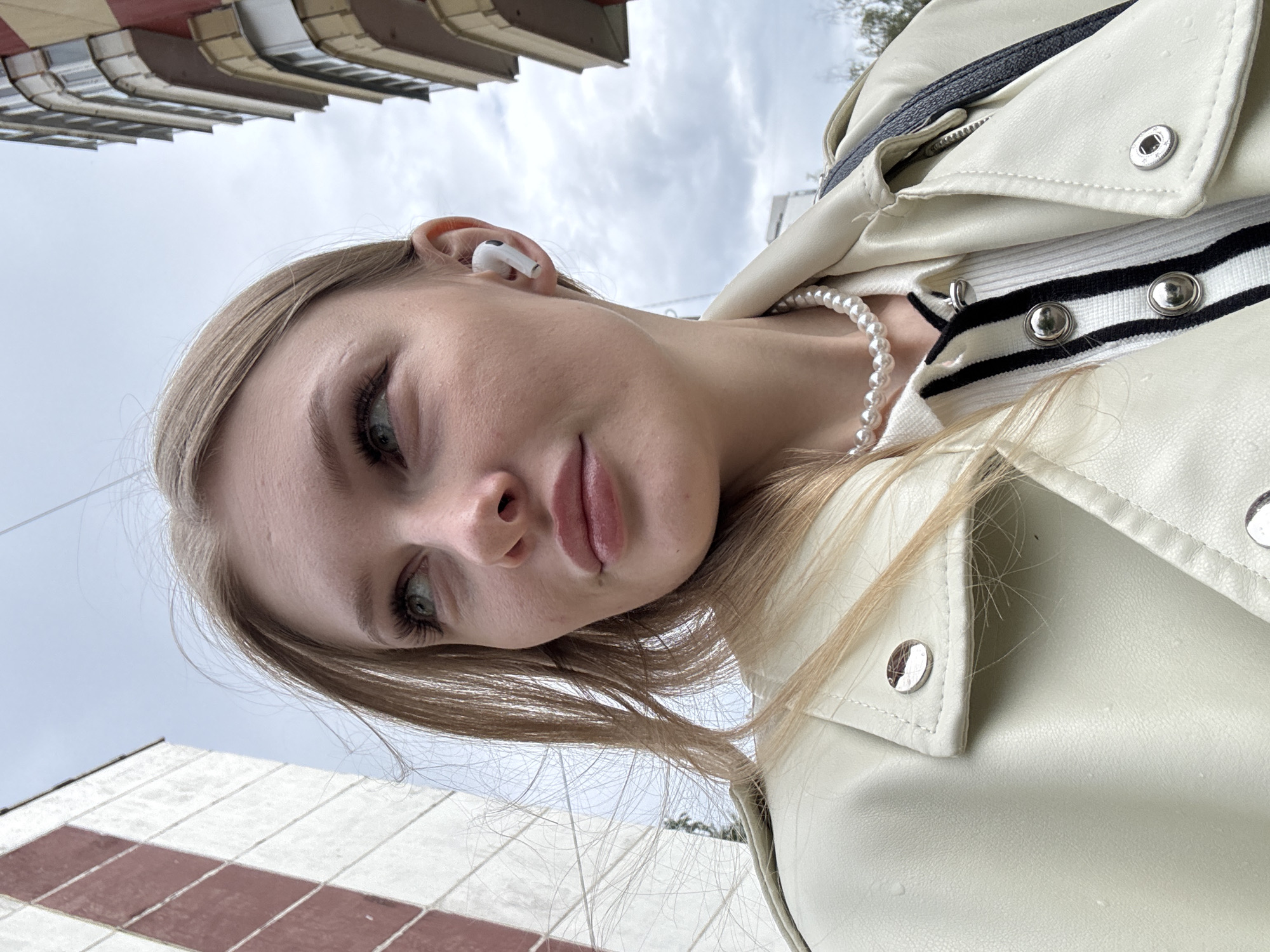The image presents a head-and-shoulders outdoor portrait of a young Caucasian woman in an urban environment, in the city of Perm, Russia, under an overcast sky. The woman, positioned at a low angle relative to the viewer, has light brown hair, light-colored eyes (appearing green/blue-green), and looks upward and to her left with a serene expression. A white wireless earbud is visibly inserted into her right ear. She wears a white pearl necklace and a light cream or off-white faux leather jacket featuring silver-toned snap buttons. Underneath the jacket, a white garment with distinct black vertical stripes and large silver circular buttons is partially visible. A dark strap, consistent with a shoulder bag, is draped over her left shoulder. The background consists of architectural elements: to the left, a tall building with a facade characterized by a checkerboard pattern of white and reddish-brown rectangular panels; to the right, a multi-story building featuring balconies and alternating sections of light yellow and reddish-brown on its exterior. No specific street names or unique landmarks are identifiable to pinpoint an exact location within Perm.