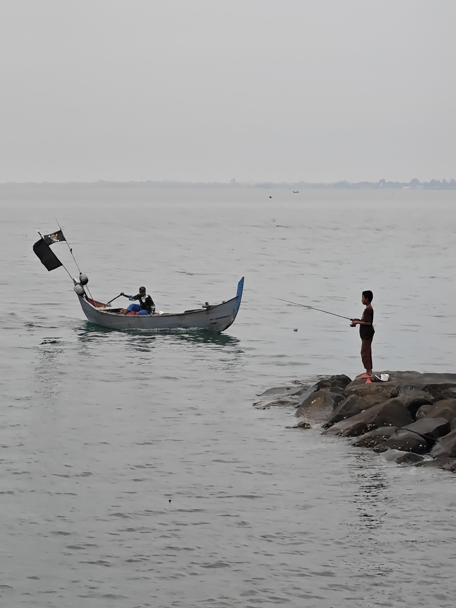 The image depicts a calm coastal scene in Lhokseumawe, Indonesia, under an overcast or hazy sky. The water is smooth and grey, with a faint, indistinct landmass visible on the distant horizon.
In the mid-ground on the left, a small, light-colored traditional boat with a blue-tipped bow is on the water. An adult, dressed in dark clothing, is seated in the boat, holding an oar or paddle, seemingly operating the vessel. Two black flags are prominently displayed at the front of the boat; one is solid black, and the other features a white skull and crossbones design. Two white buoys are also attached to the side of the boat.
On the right side of the image, a young boy stands barefoot on dark rocks that form a natural breakwater or part of the shore. He is dressed in a dark t-shirt and trousers and is actively engaged in fishing, holding a fishing rod with a reel. A small white bucket and an orange object are visible on the rocks near his feet.
The overall atmosphere is quiet and subdued, characterized by a muted color palette of grays, blues, and dark tones. The scene suggests daytime, although the lack of direct sunlight makes the exact time ambiguous. No other distinct figures or structures are visible in the immediate vicinity.