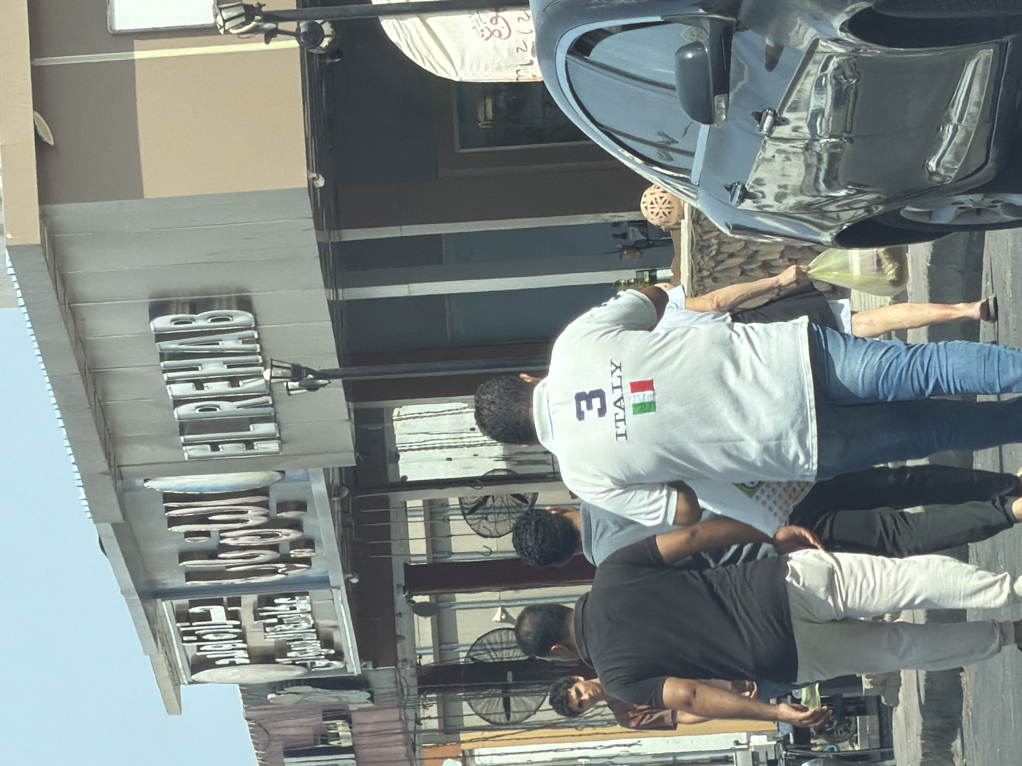 In the city of Sharm El Sheikh, Egypt, a bright, sunny day illuminates a street scene. The foreground features the rear of a dark-colored car, its polished surface reflecting the surrounding environment. Beside it, several individuals are walking. One person, in the foreground, wears a white t-shirt with "3 ITALY" printed on the back, along with the Italian flag. Another person, to their left, is dressed in a black t-shirt and light-colored trousers. Behind them, more people are visible, some walking and others standing.
The background reveals a building with prominent signage. The signage includes Arabic script and the English words "EL REHAB". The building appears to be a commercial establishment, possibly a store or restaurant, with a beige-colored facade and metal fixtures. Other elements in the background include streetlights and what appear to be large industrial fans mounted on the wall of another structure. The overall impression is of a bustling urban environment in a warm climate.