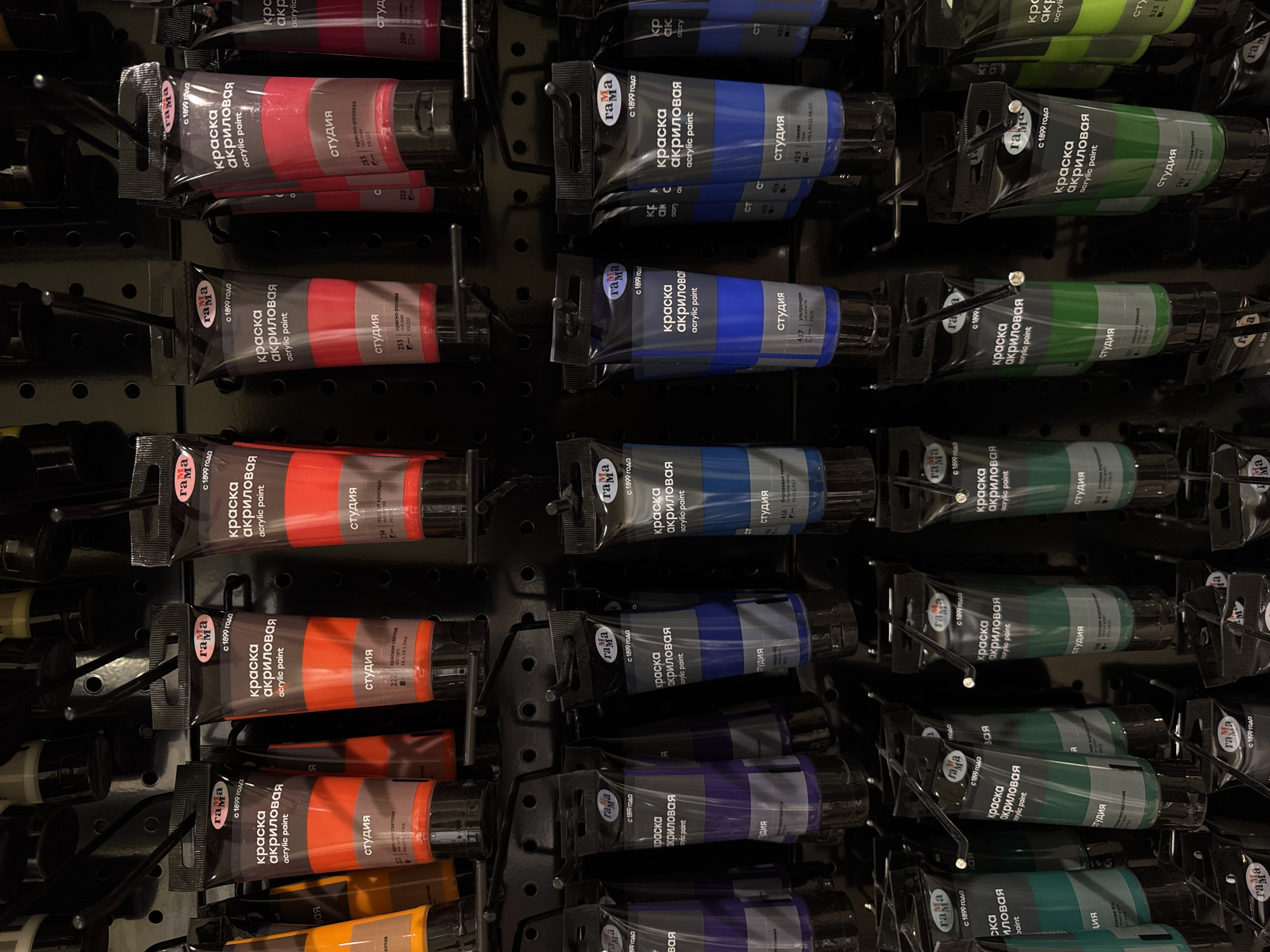 The image shows a close-up, eye-level view of tubes of acrylic paint displayed on a black pegboard in a retail setting, likely a craft store or art supply shop. The tubes are arranged in multiple rows and columns, with each tube hanging from a hook. The branding on the tubes is visible, featuring the logo "ГАМА" and text in Russian, including "краска акриловая" (acrylic paint) and "студия" (studio). Various colors of paint are visible, including orange, red, pink, purple, blue, and green. The lighting appears to be artificial, casting some shadows and highlighting the glossy surfaces of the paint tubes. The background is dark and out of focus, emphasizing the products on display. No people are visible in the image, and there are no specific geographical markers to indicate a location in Moscow, Russia.