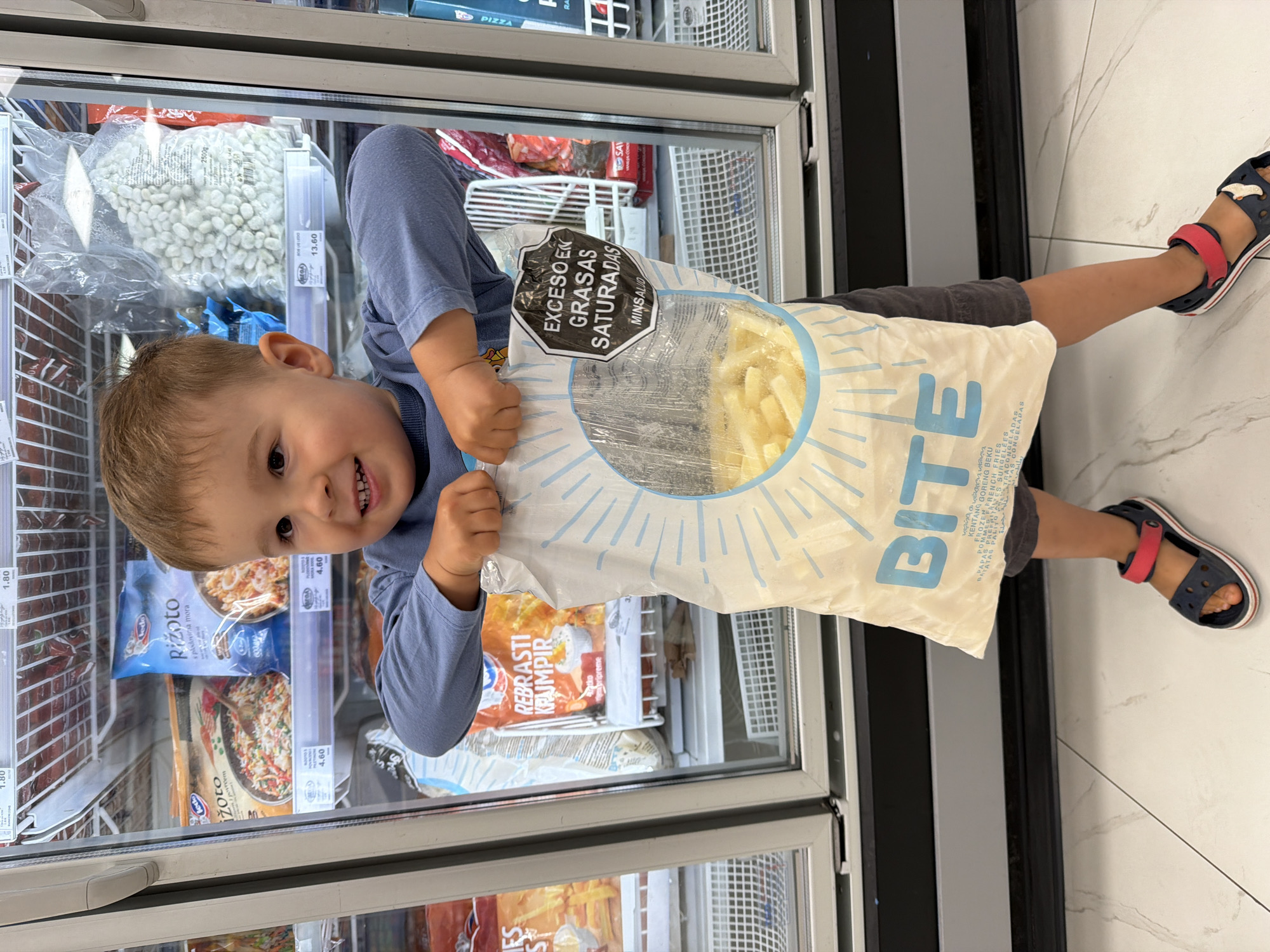 The image, rotated 90 degrees counter-clockwise, depicts a young child in the frozen food section of a grocery store or supermarket in Budva, Montenegro. The child, with light brown hair and a bright smile, looks directly at the camera. They are wearing a blue long-sleeved shirt and dark shorts, with blue and red sandals on their feet.
The main activity shows the child happily holding a large, white bag of frozen french fries labeled "BITE." The bag features a prominent black octagonal warning label stating "EXCESO EN GRASAS SATURADAS" (Excess in Saturated Fats) and "MINSALUD" (Ministry of Health), indicating a health warning system common in some Latin American countries, but also seen in other regions for imported products. Below the "BITE" logo, text in multiple languages describes the product as "KENTANG GORENG BEKU," "FRENCH FRIES," "POMMES FRITES SURGELÉES," and "PAPAS PRE-FRITAS CONGELADAS," among others. The bag is transparent in the center, revealing the frozen potato sticks inside.
Behind the child are clear glass freezer display cases, filled with various frozen food items. Visible products include bags of white beans, packages of "Rizoto" (risotto), "REBRASTI KROMPIR" (ribbed potatoes), and what appears to be frozen pizza. Price tags are visible on the shelves, showing values like "1.80," "4.60," and "13.60," along with labels such as "MEGA" and "MORA BIO." The floor is tiled with large, light-colored square tiles. The setting suggests a well-stocked and brightly lit retail environment.