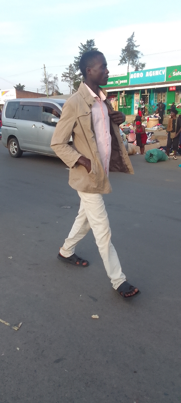 An outdoor scene in Kakunga, Kenya, depicts a street with people, buildings, and a vehicle under clear skies, likely in the late afternoon or early morning. In the foreground, a man of dark complexion is walking towards the right, his body angled slightly towards the camera. He wears a light brown jacket with his right hand in its pocket, a pink collared shirt, white trousers, and dark sandals. His left hand is seen adjusting or holding his jacket.
In the mid-ground on the left, a silver minivan is either parked or slowly moving. Behind the man and to the right, several other individuals are visible, some gathered around what appears to be goods or clothing laid out on the ground, and a large green bag rests nearby.
The background features commercial buildings. A prominent green building displays various texts, including "AGRO AGENCY," "m_pesa," "Safaricom," and "FARM INPUTS," suggesting businesses related to agriculture or mobile money services. Trees are scattered behind these structures. The road surface is asphalt, with some minor debris visible. The overall activity suggests a bustling street or market environment. No strong emotions are discernible from the individuals.
