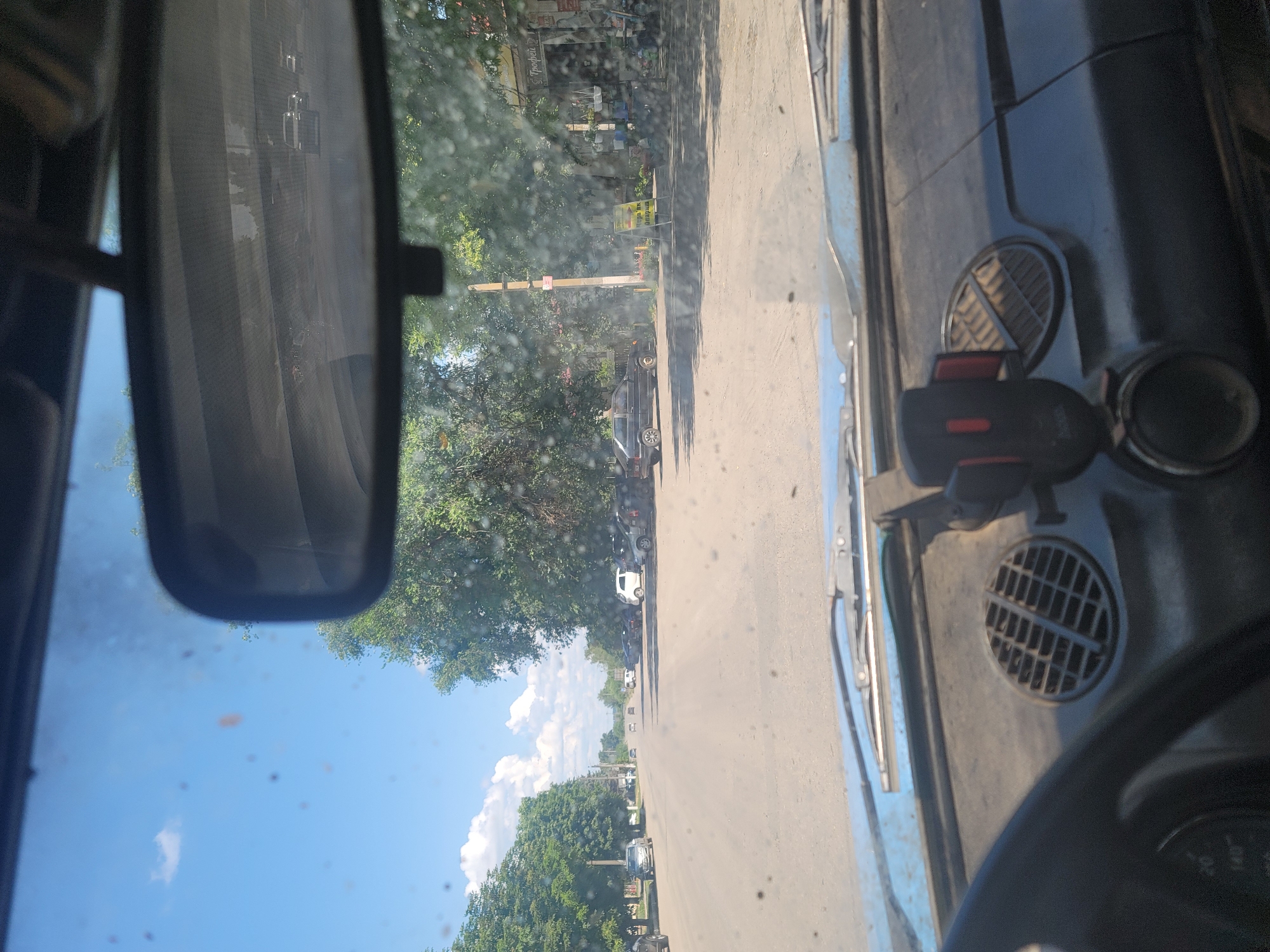 The image, taken from the interior of a car, provides a view of a street in Pereshchepyne, Ukraine. The perspective is from the driver's seat, looking forward through a windshield speckled with dust and water droplets. On the dashboard, a black cell phone holder with a red accent is visible to the right of the steering wheel. The street itself is paved and appears to be a two-lane road with parked cars on both sides. On the left side of the road, there are houses and trees lining the street. On the right side, there are more trees and a building with a sign that reads "Трофей" (Trophy) in Cyrillic script, suggesting a shop. A yellow advertising stand is also visible near the building. The sky is blue with scattered white clouds, and the overall scene suggests a bright, sunny day. Several vehicles are parked or driving on the street, including a white car, several dark-colored sedans, and a black hatchback. The car from which the photo is taken is a light blue vintage model.