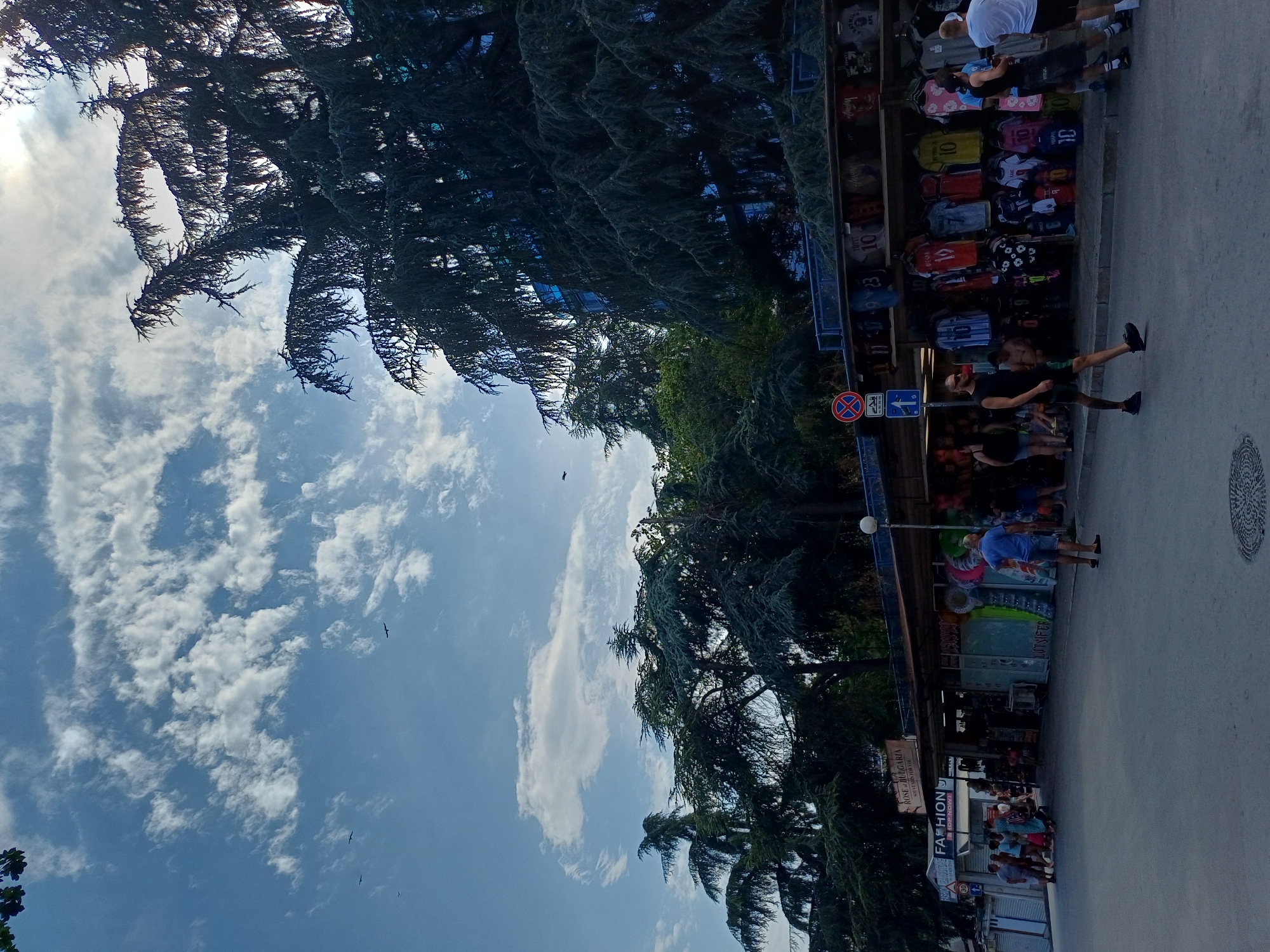 In Varna, Bulgaria, along a waterfront promenade lined with large evergreen trees, a street-level view captures a bustling market area. To the right, several market stalls display an array of colorful t-shirts, many featuring sports team logos or numbers. A man in a black t-shirt and shorts walks across the pavement, while a man in a blue t-shirt and denim shorts stands facing away from the viewer, towards the stalls. A group of people, including men, women, and children, are gathered near a stall labeled "FASHION," suggesting a tourist-oriented shopping district. The sky above is a vibrant blue, dotted with scattered white clouds, and illuminated by the bright sun, casting a slight glare. The overall scene depicts a lively summer day in a popular Varna locale.