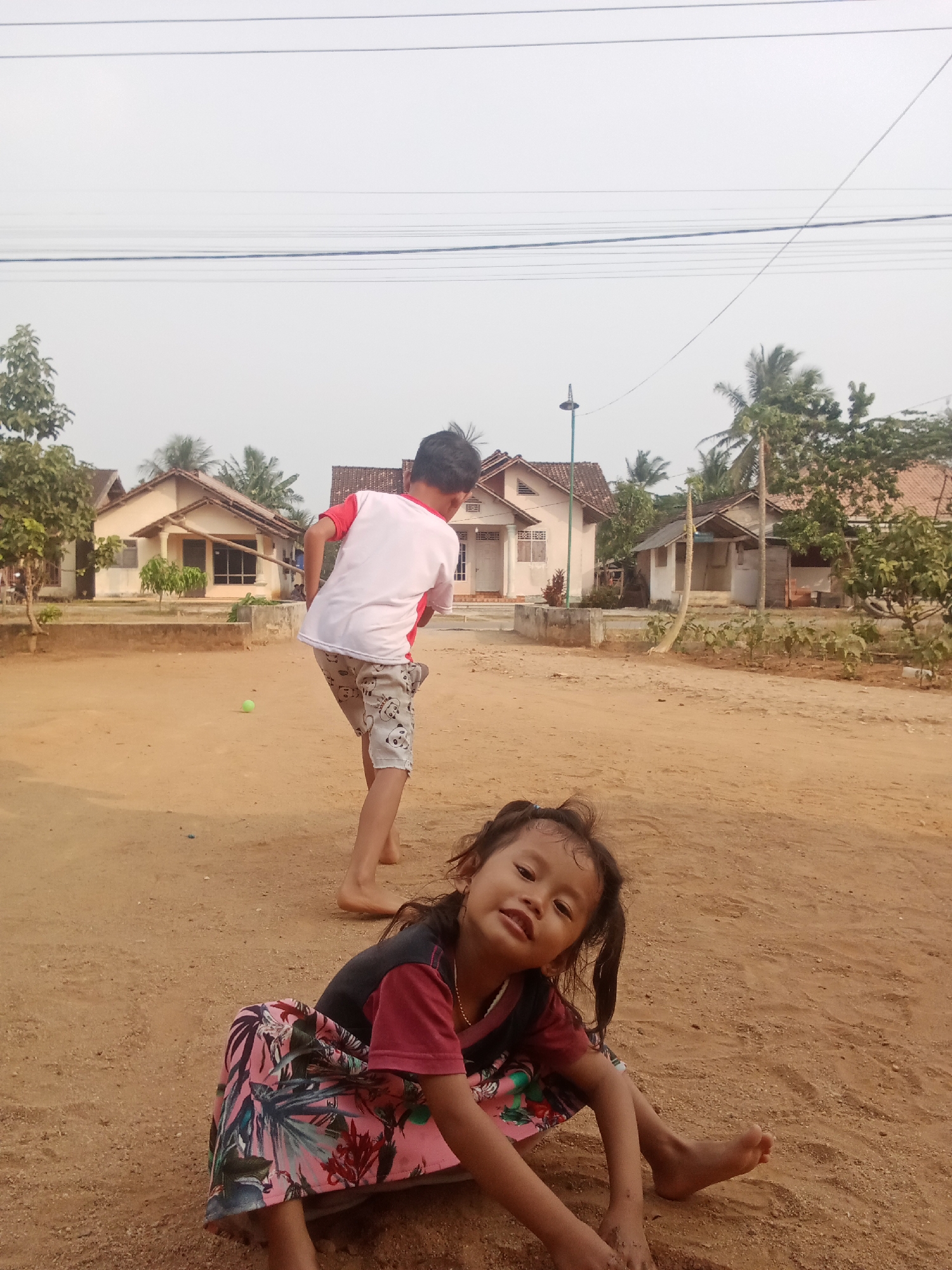 In a dusty, unpaved outdoor area that appears to be a village setting in Unknown, Indonesia, two young children are present. A young boy, facing away from the camera and to the left, is in the process of throwing a green ball. He is wearing a white t-shirt with red accents on the sleeves and collar, and light-colored shorts patterned with small cartoon-like figures. In the foreground, a young girl is sitting on the ground, facing the camera. She has dark hair styled in pigtails and is wearing a dark shirt with red sleeves and a pink skirt with a floral pattern. She is barefoot. Behind the children, several modest houses with terracotta roofs are visible, along with scattered trees and utility poles with overhead wires. The ground is primarily composed of dry, sandy soil, indicating a rural or semi-rural environment. The overall lighting suggests it is daytime, with the sky appearing hazy.