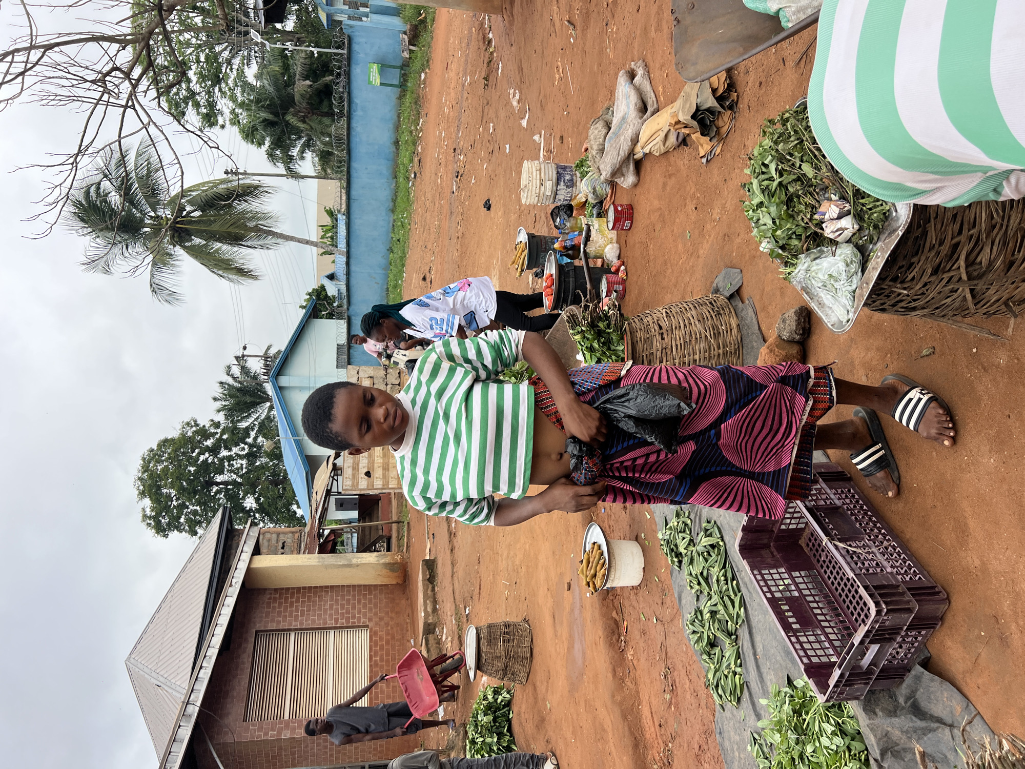In Nkpor, Nigeria, an outdoor market scene unfolds under an overcast sky. A young woman with a short haircut, wearing a green and white horizontally striped cropped t-shirt and a colorful patterned skirt, stands center frame. She is holding a black plastic bag in her hands, which are partially obscured by her skirt. Her feet are adorned with black and white striped sandals. In front of her, a purple plastic crate is placed on a black tarpaulin, surrounded by green leafy vegetables. To her right, a woven basket overflows with more green produce, and further to the right, a pile of goods wrapped in burlap sacks and a wheelbarrow are visible. Behind her, a variety of items are spread out on the ground, including tomatoes, corn on the cob, cans, and buckets. To the left, a brick building with a corrugated metal roof stands alongside other structures. Two men are seen in the background; one is pushing a red wheelbarrow, and another, in a gray tank top, is walking away from the camera. A woman on a motorcycle, wearing a white t-shirt with graphics, is also present in the mid-ground. The ground is covered with reddish-brown soil, characteristic of the local terrain. The overall environment suggests a bustling marketplace.