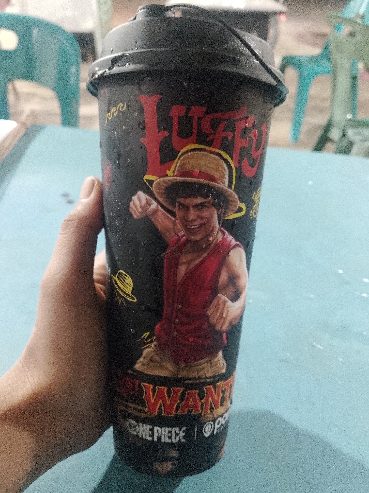 A hand holds a black, plastic cup with a lid. The cup is decorated with an image of a live-action version of the character Luffy from the anime One Piece. Luffy is wearing his signature straw hat and red vest. The cup also has the words "Luffy" and "Most Wanted" printed on it. The cup is covered in condensation.
The background is blurry, but it appears to be an outdoor setting with blue and green plastic chairs. The location is Lhokseumawe, Indonesia.