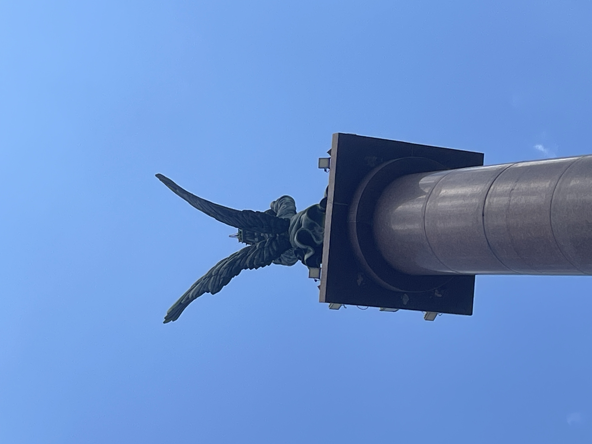 An upward shot of a bronze statue of an angel with outstretched wings atop a granite column against a clear blue sky. The statue appears to be a monument. The column is cylindrical and a rich brown color, with distinct horizontal banding. At the top of the column, a dark, square pedestal supports the statue. Several small, square lights are visible on the edges of the pedestal. The scene is brightly lit, suggesting daylight. This statue is located on Malaya Zemlya in Novorossiysk, Russia.