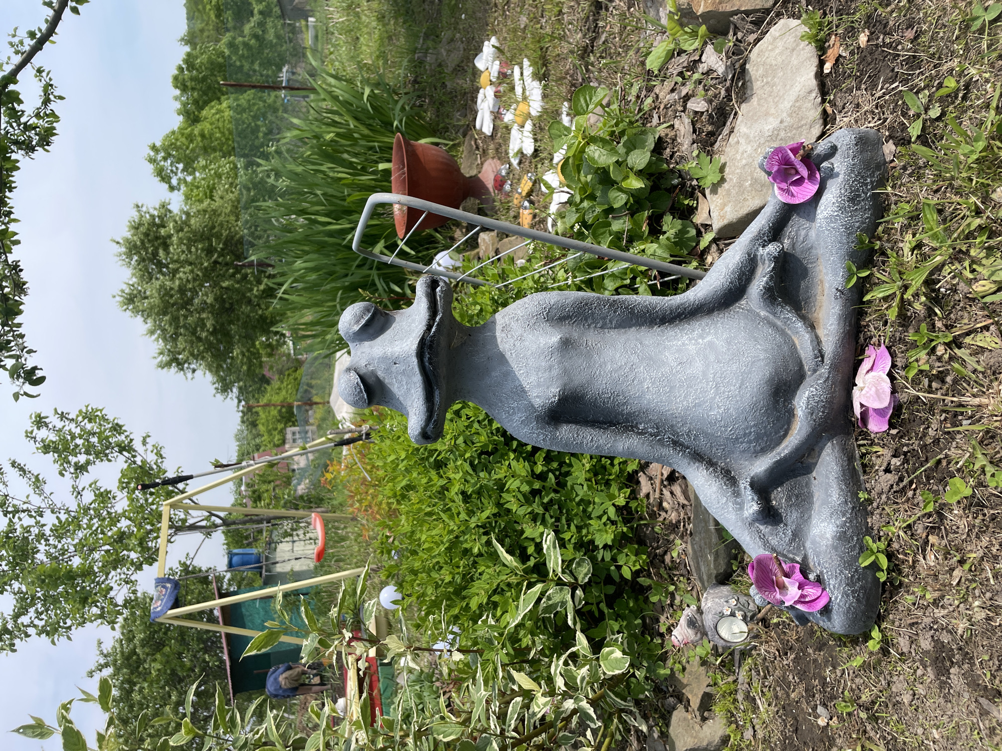 A gray, stylized statue of an animal, possibly a frog or hippo, lies horizontally in a garden. Purple flowers are placed on the statue. Around the statue, there are various green plants and white daisies. In the background, there is a swing set and other garden decorations, including a brown pot and a metal rack. The sky is overcast. The scene appears to be in a yard or garden. Given the inclusion of Yasnoe, Russia, in the instructions, this location is assumed.