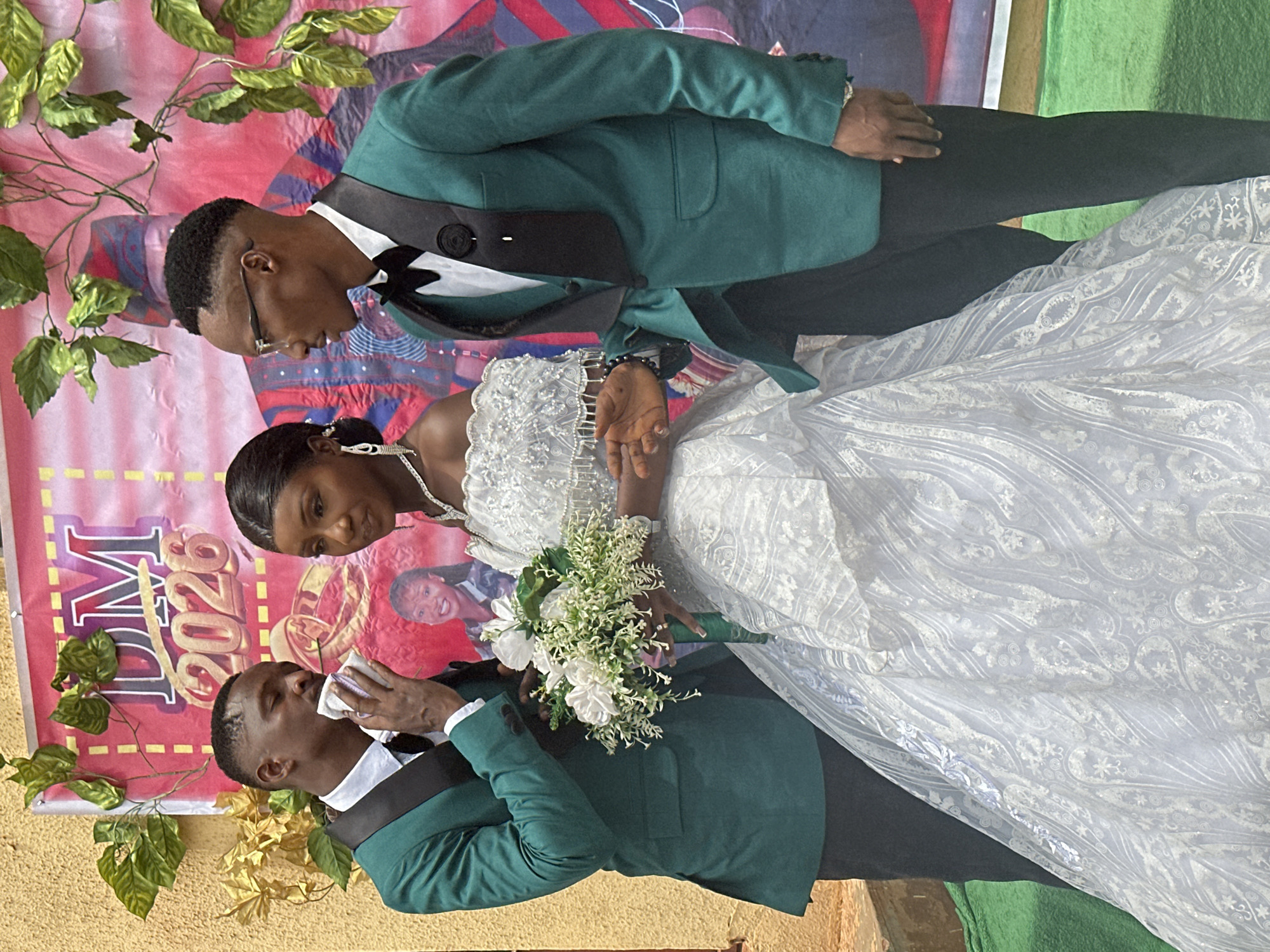 A wedding photo of a bride and groom and an attending guest is captured in Unknown, Unknown. The bride is adorned in a detailed white wedding gown, and she holds a bouquet of white flowers. Two men wearing green suits with black lapels frame her. One of them is wiping his face with a white cloth, while the other looks down towards the bride. Behind them is a banner with the lettering "DM 2026" on a red background with gold accents. Decorations of green and yellow vines adorn the backdrop. The ground appears to be tan or brown, and a green cloth is visible in the bottom right corner.