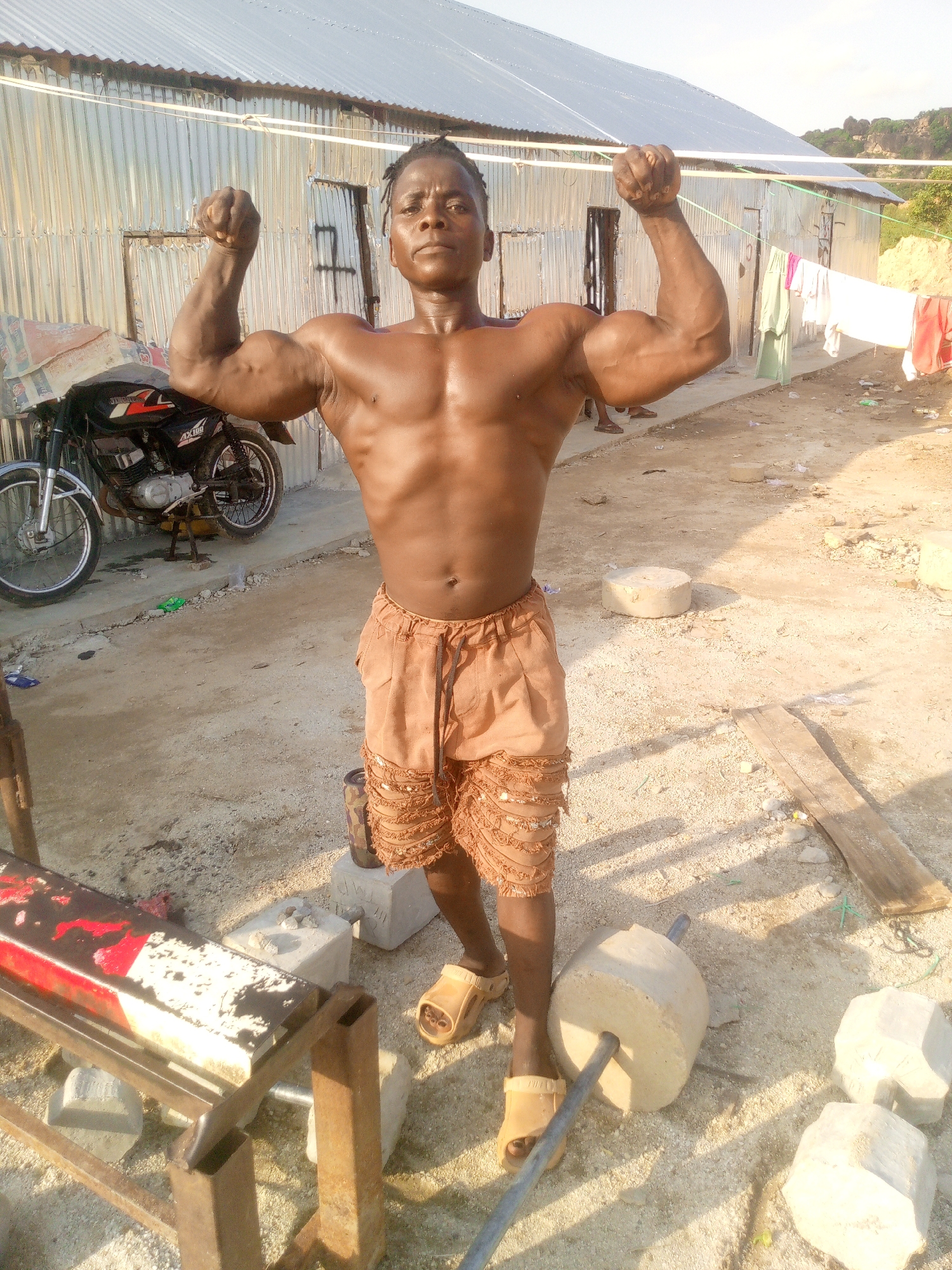 A muscular Black man is posing in front of a corrugated metal building in Indo, Nigeria. He is flexing his biceps, and his skin is glistening with sweat. He is wearing light brown shorts and tan sandals. To his left, a motorcycle is parked. In the background, laundry hangs on a clothesline. The ground is dirt and gravel, and there are several concrete weights scattered around. The building appears to be a simple, rustic structure, possibly a workshop or storage area. The sky is bright and clear, suggesting a sunny day.