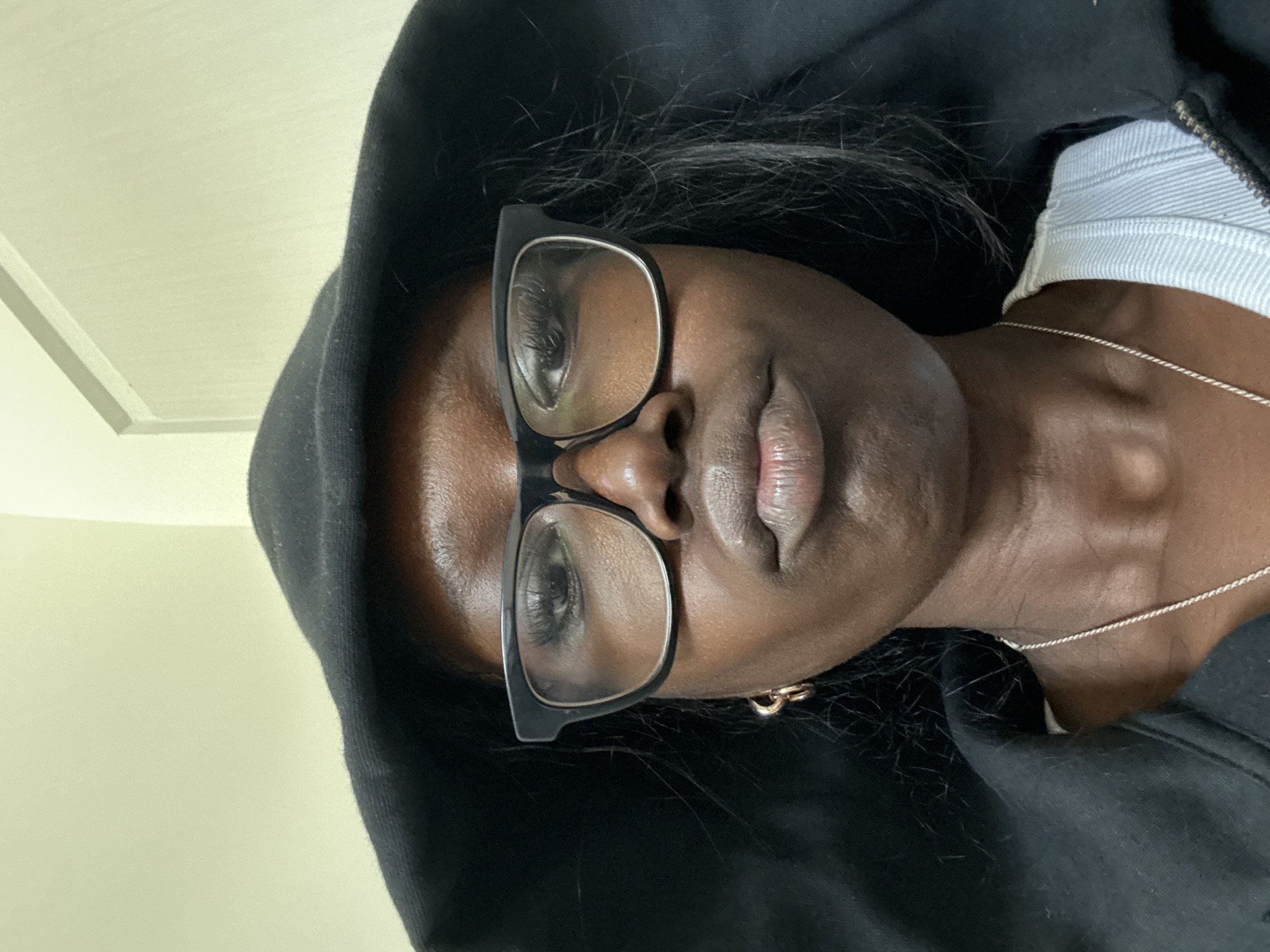 A person wearing glasses and a black hooded sweatshirt is facing the camera. They also wear a white top with a zipper, and a silver necklace. They are indoors. The location is unknown.