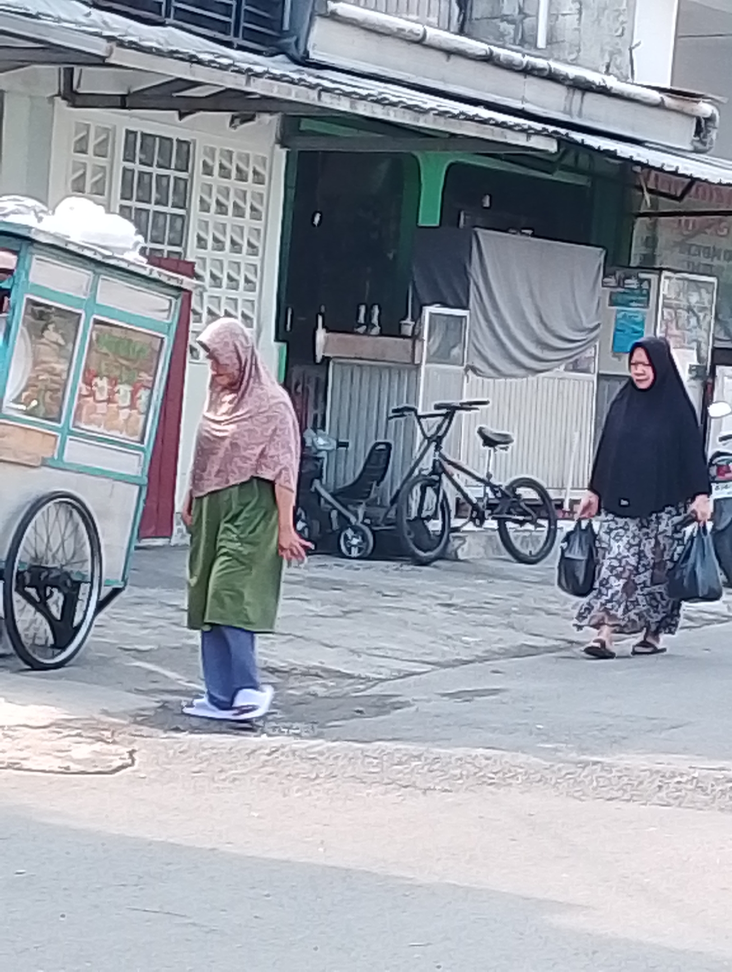 This is a full-shot, eye-level, outdoor photograph of two women walking on a paved sidewalk in front of a building in Bogor, Indonesia. The woman on the left is wearing a light pink hijab, a green skirt, and blue pants, with white shoes. She is standing next to a food cart. The food cart is turquoise and white with a display window showing images. The woman on the right is wearing a black hijab and a patterned dress, with dark sandals. She is carrying two black bags. Behind the women, there are bicycles and a stroller. The building in the background appears to be a shop with large windows and awnings. The scene is lit by natural sunlight, creating shadows on the ground.