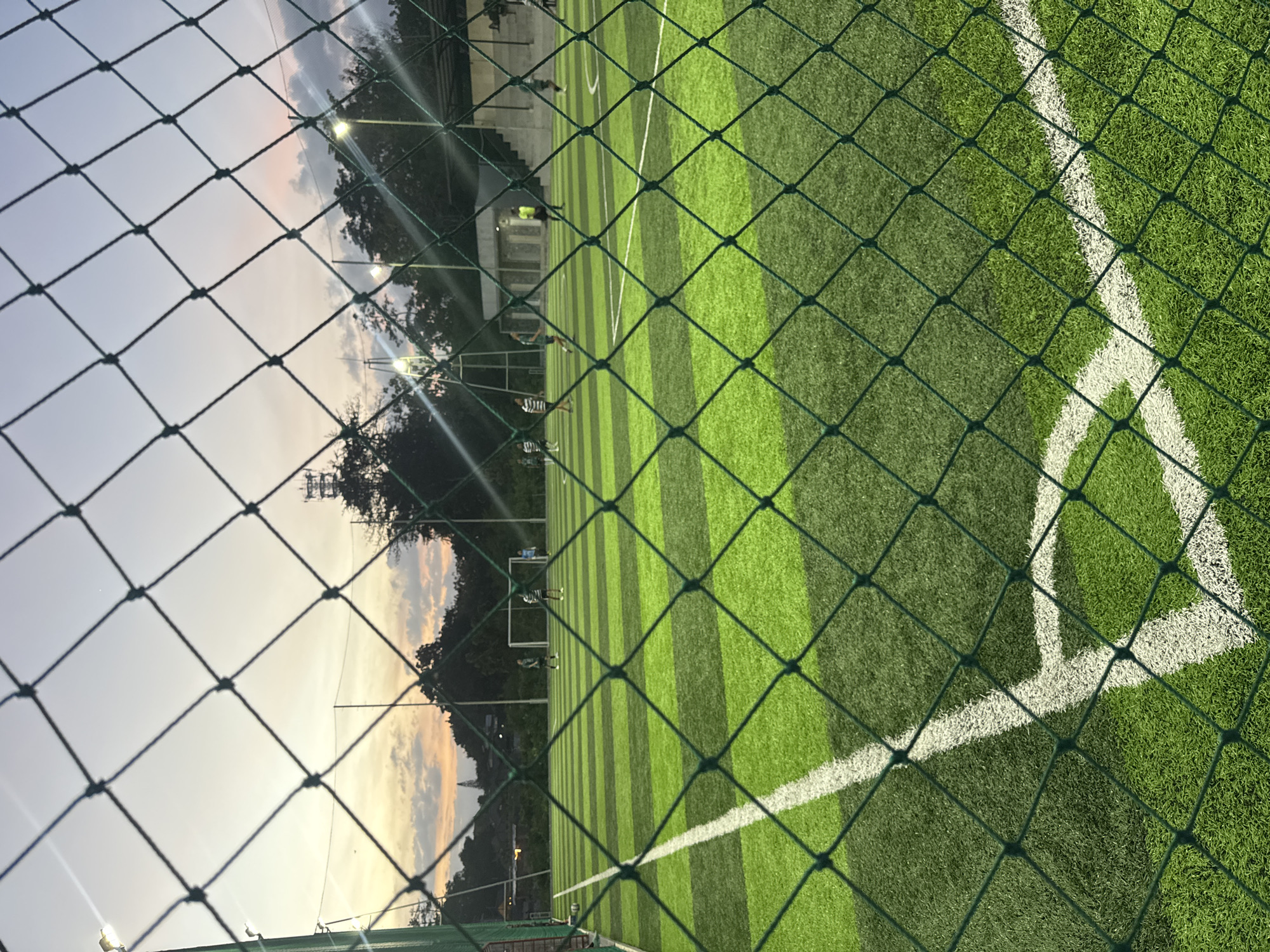 The image captures a soccer field, framed by a green chain-link fence. The field has artificial turf with alternating light and dark green stripes. White lines mark the boundaries and corners of the field. In the background, trees and a structure are visible. The sky suggests it is either dawn or dusk. The location is Kuta Selatan, Indonesia.