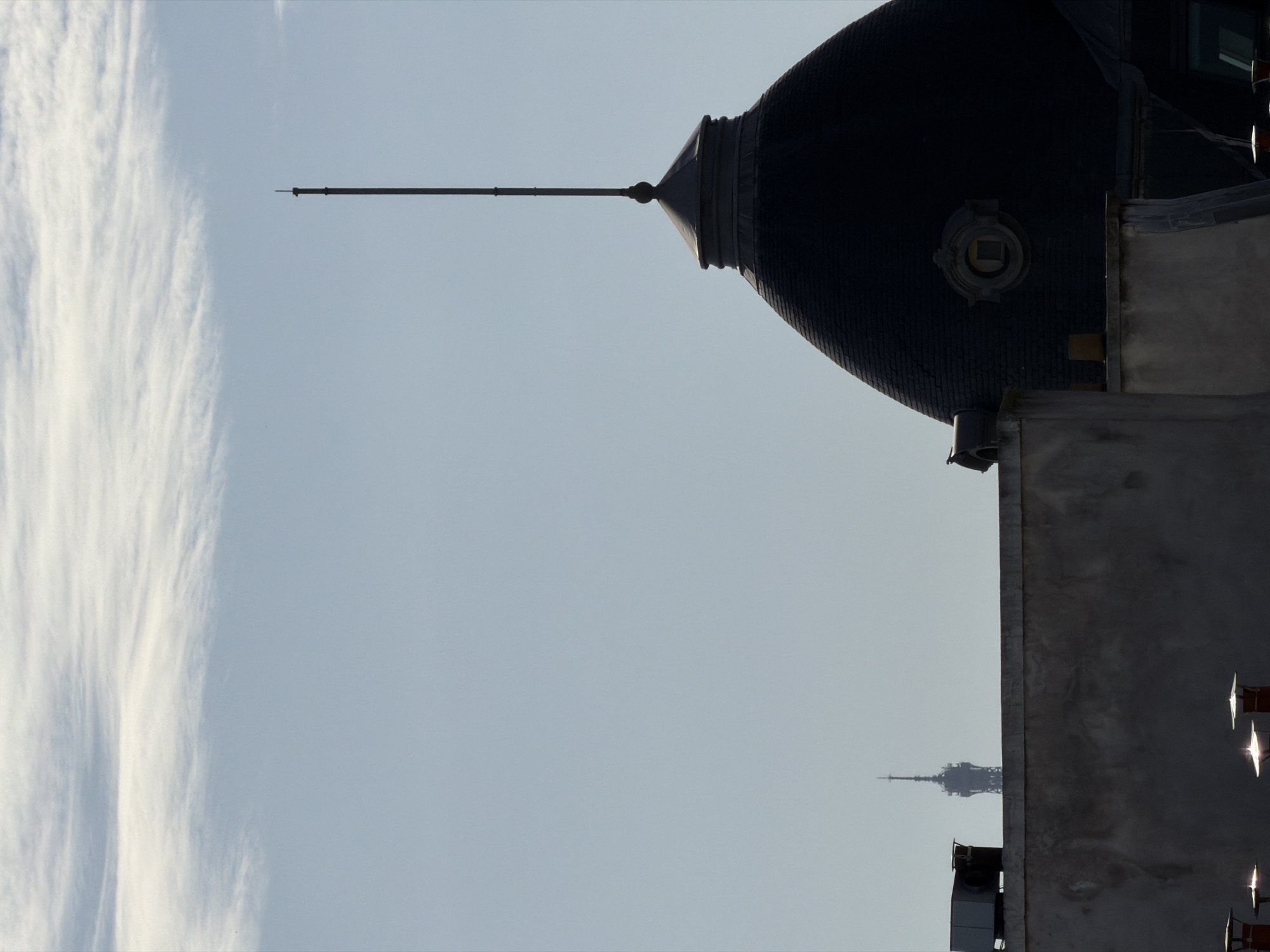 The image displays an urban sky scene with architectural elements. A large, dark, textured dome with a slender spire pointing left dominates the upper right portion of the frame. The dome, possibly covered in slate or metal tiles, features a small, ornate, rectangular detail on its side. Below and to the right of the dome, parts of a building structure, likely a rooftop, are visible in dark tones.
On the far left, a vertical band of wispy, white clouds stretches against a pale blue sky, which occupies most of the central and left-hand areas of the image. In the lower right background, the unmistakable silhouette of the Eiffel Tower is faintly discernible against the bright sky, confirming the location as Paris, France. Some bright, reflective glints are visible on the edge of the building structure in the bottom right, suggesting sunlight catching metallic or glass surfaces.
The scene depicts a static daytime view. The bright sky indicates daylight, but the deep silhouette of the dome and foreground buildings suggests the sun is positioned behind them or at an angle that creates strong backlighting. There is no visible activity or text. The overall impression is one of iconic Parisian architecture against a serene sky.