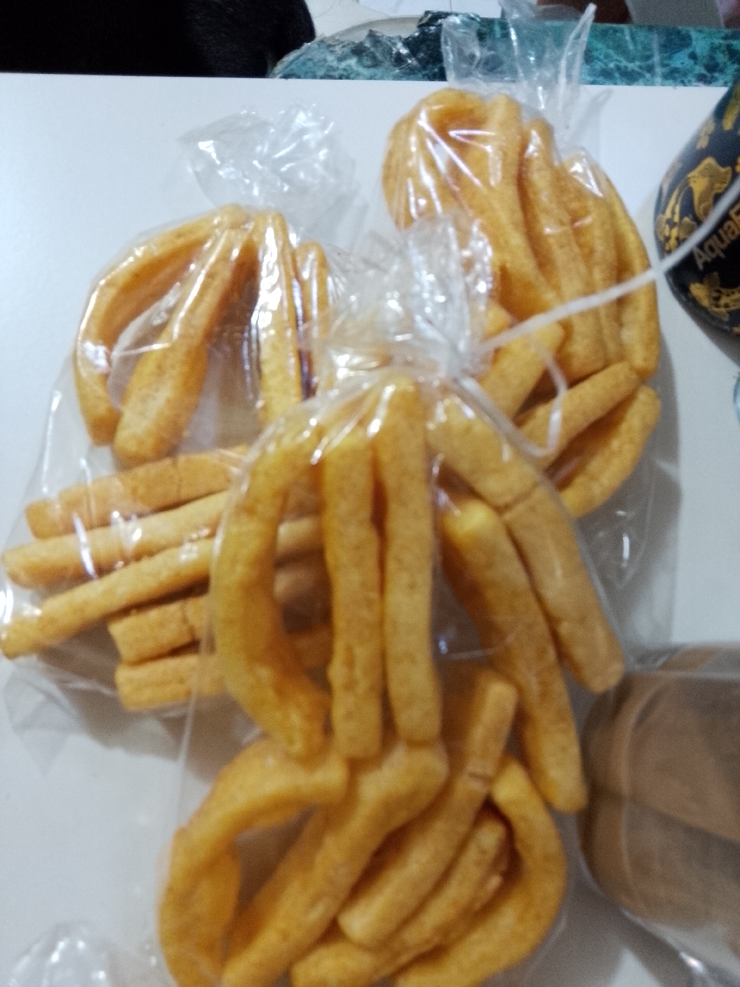 An indoor scene, situated in Davao City, Philippines, displays five transparent plastic bags filled with deep-fried snacks, resting on a flat white surface. Three visible bags predominantly contain numerous golden-yellow, crescent-shaped, puffy fried snacks, while one bag is filled with straight, golden-yellow, rectangular fried sticks. A fifth bag, containing the crescent-shaped snacks, is partially visible at the bottom. The snacks maintain a uniform golden-yellow color. The white surface serves as the foreground. In the upper left background, a dark, out-of-focus, potentially furry object is present. To the upper right, a mottled green and white (or blue) patterned countertop, consistent with marble or granite, is visible. A black cylindrical container with gold embellishments, bearing the partial inscription "AquaF" (likely AquaFlask), is situated on the right side of the background. A brown, indistinct object is partially visible in the bottom right corner. No individuals or active interactions are observed.