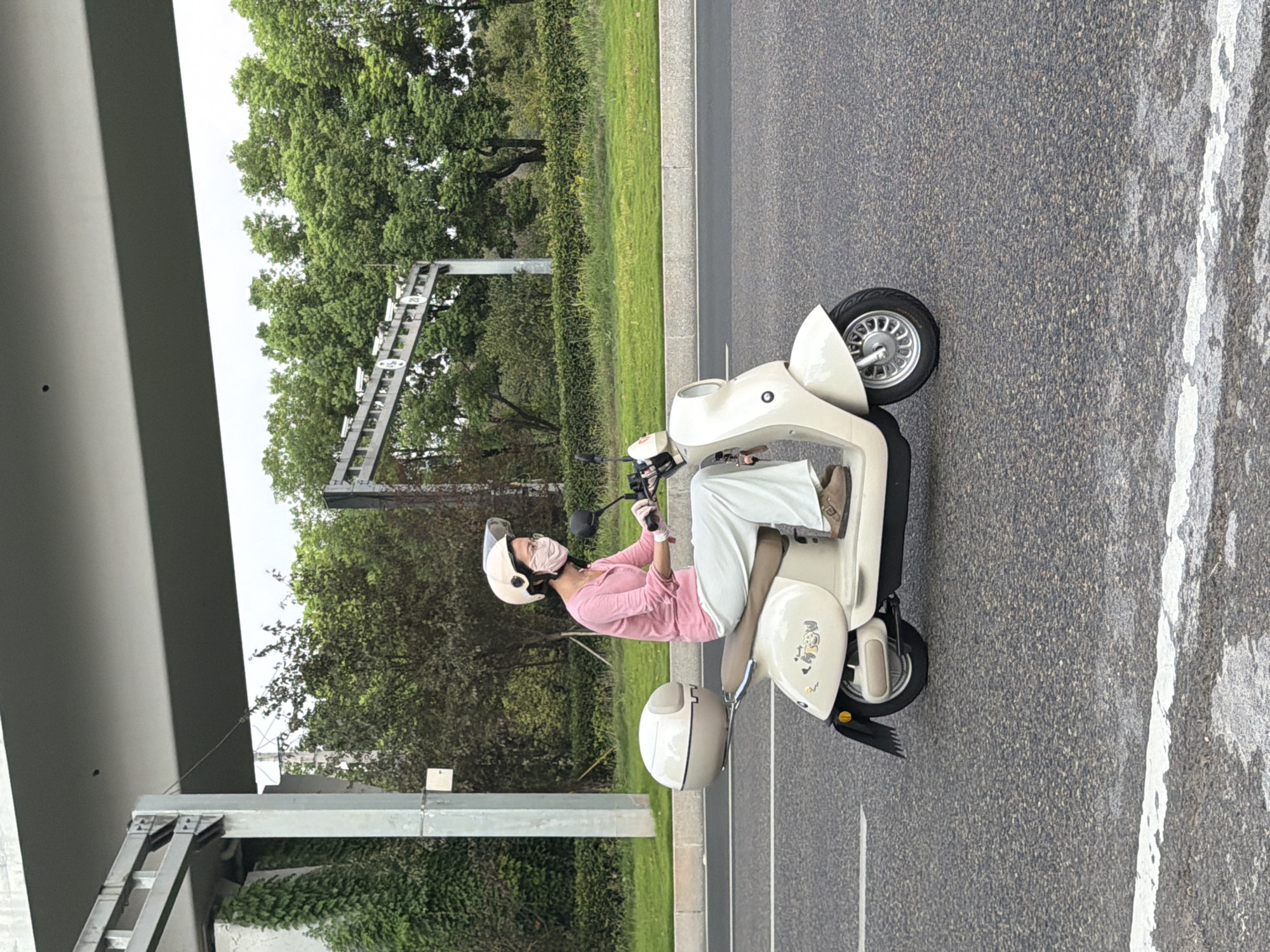 A woman wearing a pink long-sleeved shirt, white pants, a white helmet, and a pink face mask rides a cream-colored scooter on an asphalt road. She is wearing glasses and white gloves. The scooter has a rounded rear storage compartment and decorative decals on its side. In the background, there is a green grassy area with trees and a grey overpass structure with lights. The scene appears to be in a city in China, potentially Jiangjin, given the context.