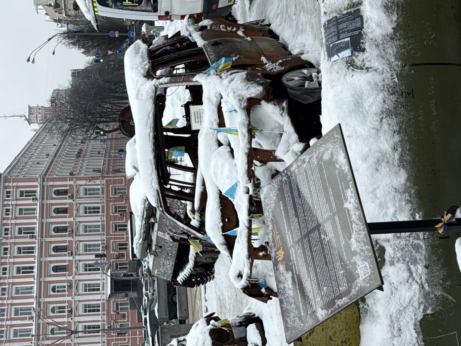 This image depicts an outdoor exhibition of destroyed military equipment on Khreshchatyk Street in Kyiv, Ukraine, under a layer of snow. In the foreground, a dark-colored informational plaque on a metal stand is angled upward, partially covered in snow. It features the Ukrainian trident emblem and text in Ukrainian and English, identifying the display as an "Exhibition of destroyed Russian military equipment." A small blue and yellow ribbon is tied to the stand's base.
Immediately behind the plaque, a heavily rusted, burnt-out vehicle frame, consistent with a large car or SUV, is prominently displayed. Its roof, doors, and interior components are largely absent, and its entire structure is covered in white snow. Numerous small Ukrainian flags (blue and yellow) are affixed to the remaining metal frame.
Behind this vehicle, partially obscured, is a snow-covered tracked armored personnel carrier (APC), also exhibiting damage and adorned with small Ukrainian flags. To the far right, a portion of a white utility vehicle or ambulance, also snow-covered, is visible.
In the background, a large, multi-story building with a distinctive pink facade and numerous white-framed windows lines the left side of the street. Further down the street, bare trees, lampposts, and traffic lights (one showing green) are visible, along with a building featuring a prominent spire. The entire ground surface is covered in a uniform layer of snow, indicative of a winter setting. No individuals are visible in the scene.