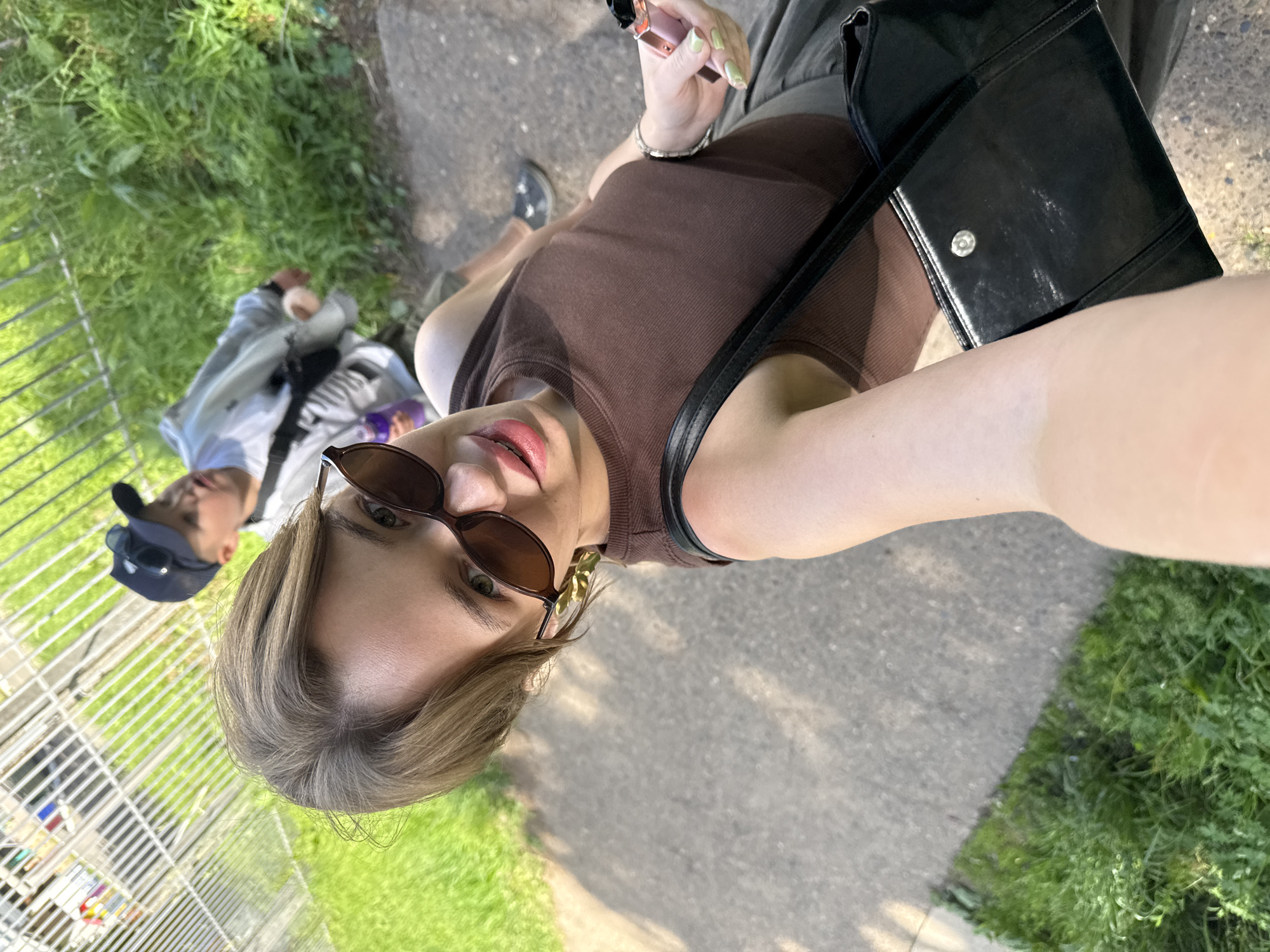 A young woman with short, light brown hair and sunglasses is taking a selfie outdoors. She is wearing a brown sleeveless top and a grey skirt. She is holding a pink vape pen in her right hand. A black purse with a silver clasp is slung over her left shoulder. In the background, a young boy wearing a blue baseball cap and a grey jacket is walking away from the camera. They appear to be on a sidewalk next to a grassy area and a metal fence. The image was taken in Chaikovsky, Russia.