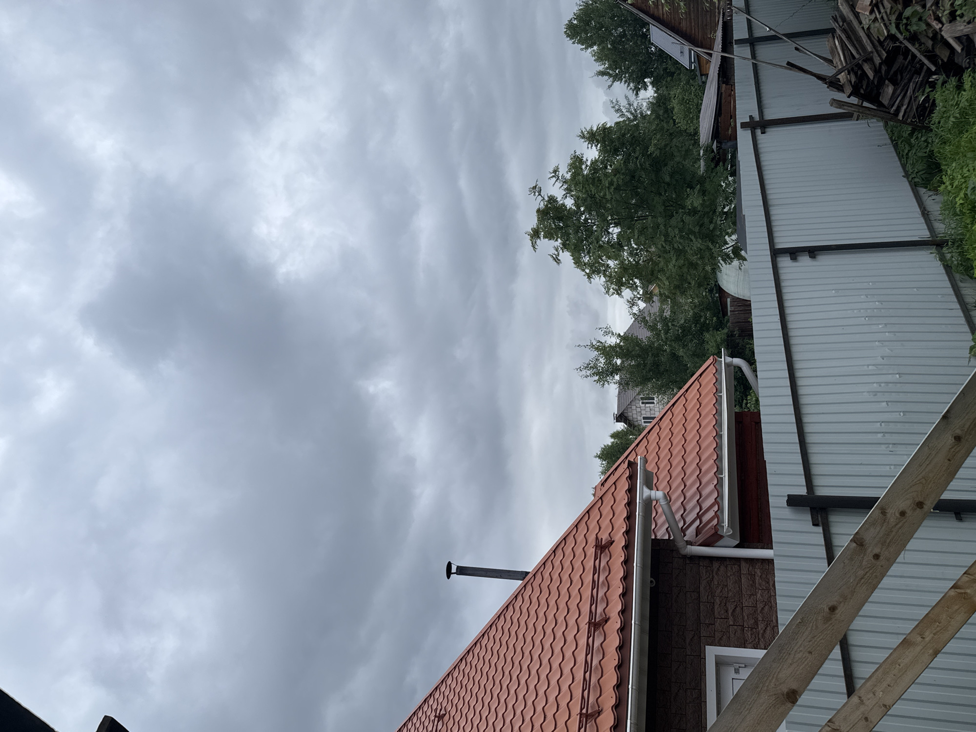 This is a vertically oriented, close-up shot of a residential building's exterior, with an overcast sky dominating the upper portion of the frame. The scene appears to be in a rural or suburban setting, likely in Perm, Russia, given the context.
The main subject in the foreground is a red-tiled roof with a visible chimney and a white gutter system. The building beneath the roof has a textured brown facade, possibly brick or siding, and a white-framed window. To the right of this building, a light gray corrugated metal fence runs horizontally across the mid-ground, supported by dark metal posts. Behind the fence, a stack of weathered wooden planks is piled high, suggesting construction or renovation work. Trees with dense green foliage are visible behind the fence and the first building, with glimpses of other structures and more trees further in the distance.
The sky is filled with heavy, dark gray clouds, indicating a very overcast day, possibly on the verge of rain. There is no visible text in the image. The overall mood is somewhat somber and subdued due to the weather. The scene captures a moment of everyday life, possibly focusing on the architecture and the surrounding environment of a home.