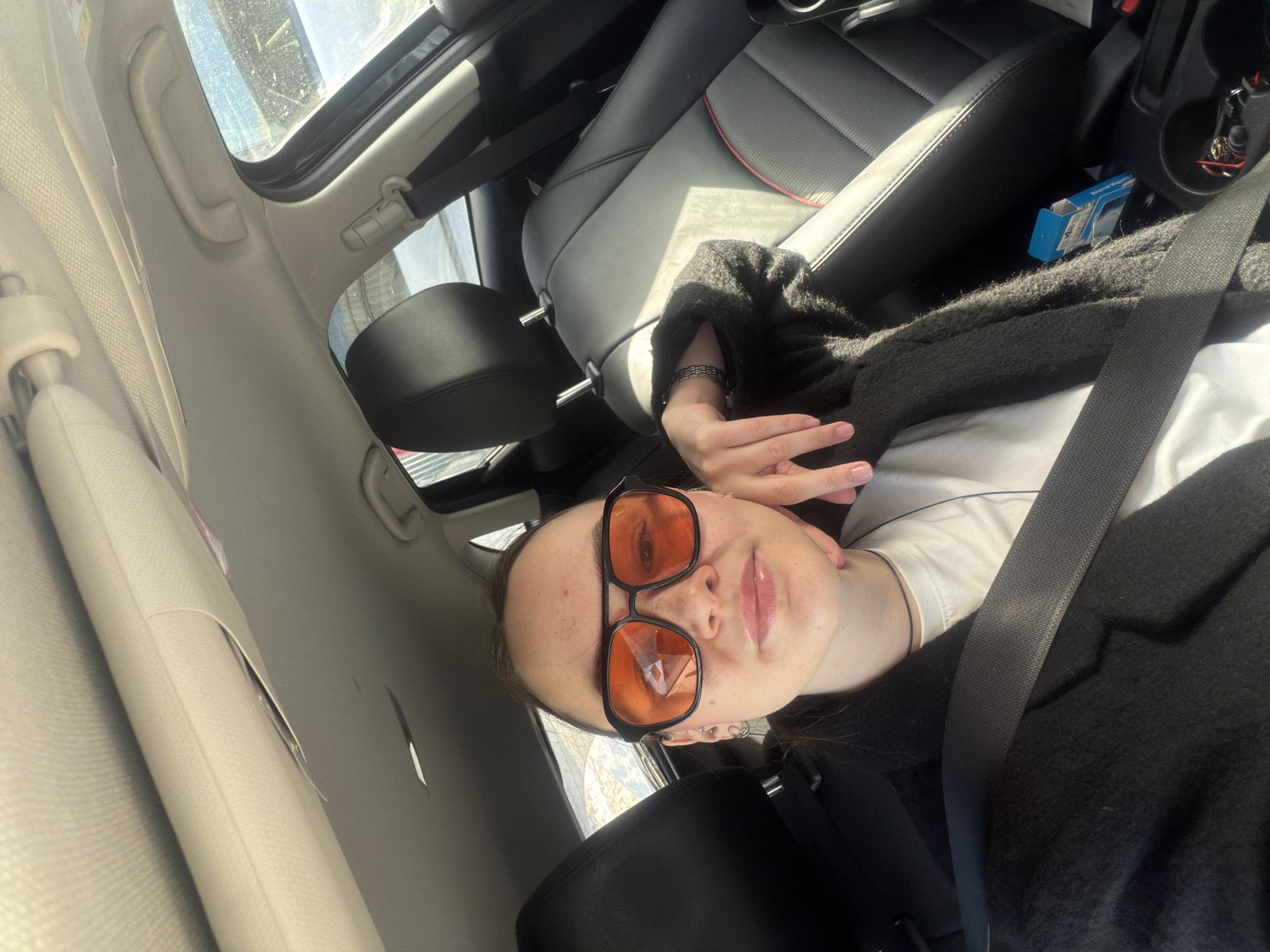 A young adult female is depicted seated in the driver's position inside a vehicle in Chaikovsky, Russia. She wears large, square-framed sunglasses with orange-tinted lenses, a black choker, a white t-shirt, and a black textured cardigan. Her dark hair is pulled back, and she has visible freckles. Two small hoop earrings and a stud are present on her left ear. A dark, multi-strand bracelet adorns her left wrist, and her fingernails display light pink polish. She is secured by a black seatbelt across her chest and shoulder, with her left hand resting against her chin. The car interior features black leather seats with red stitching accents. The passenger seat is unoccupied. The light beige headliner above shows visible peeling or tearing near the sun visor. Sunlight streams into the vehicle from the passenger side, illuminating the interior. A blue box and a cup holder containing wired earbuds are visible in the center console area. Through the windows, a snow-covered exterior environment is discernable.