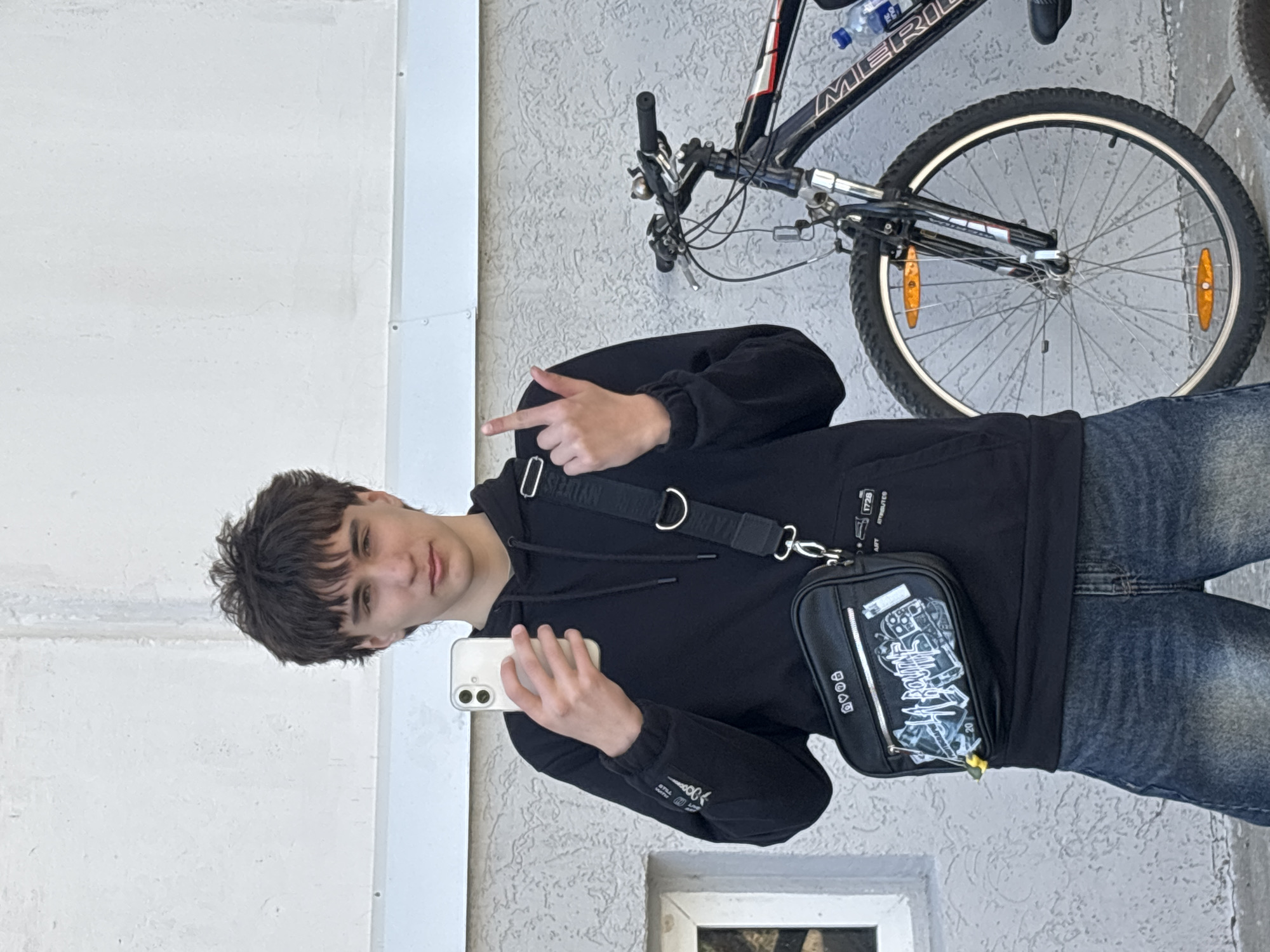 A young man is standing in front of a light gray textured wall with a white panel on the left and a window near the bottom right, with a bicycle partially visible at the top right. The man is wearing a black hooded sweatshirt and jeans. He is holding a white smartphone and taking a selfie. He is pointing his left index finger towards the upper right. The location is specified as Chaikovsky, Russia.