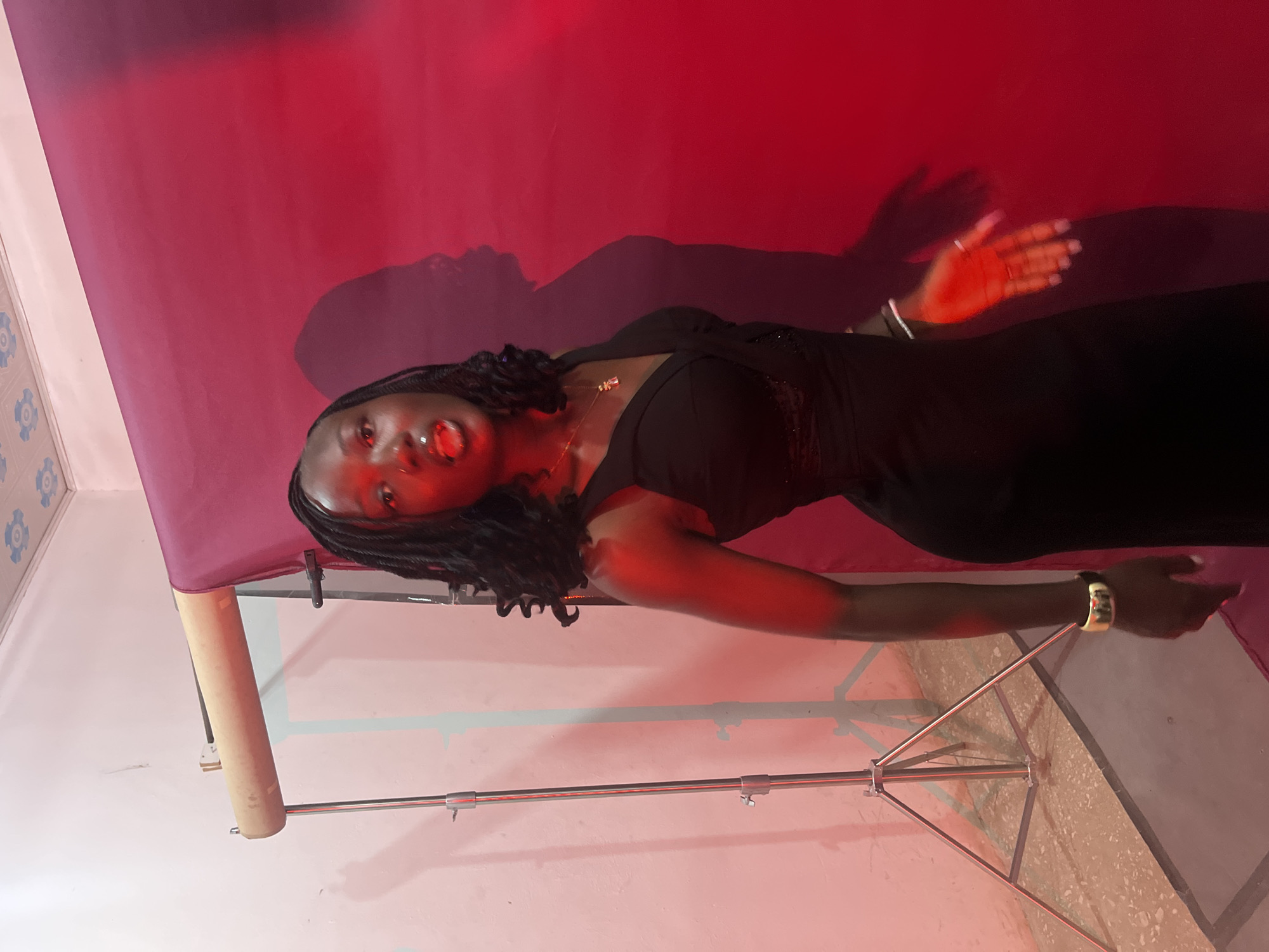 A young Black woman with dark, braided hair stands in front of a red backdrop, illuminated by red lighting. She is wearing a black dress with a V-neckline and a lace detail at the bust. A gold necklace hangs around her neck. On her left wrist, she wears a wide gold bracelet, and on her right hand, a thin silver bracelet and a ring are visible. She is looking towards the camera with her mouth slightly open. To the left of the woman, a photography backdrop stand with a light-colored fabric is visible, casting shadows on a white wall. The floor appears to be tiled. The scene is likely a studio setup for photography.