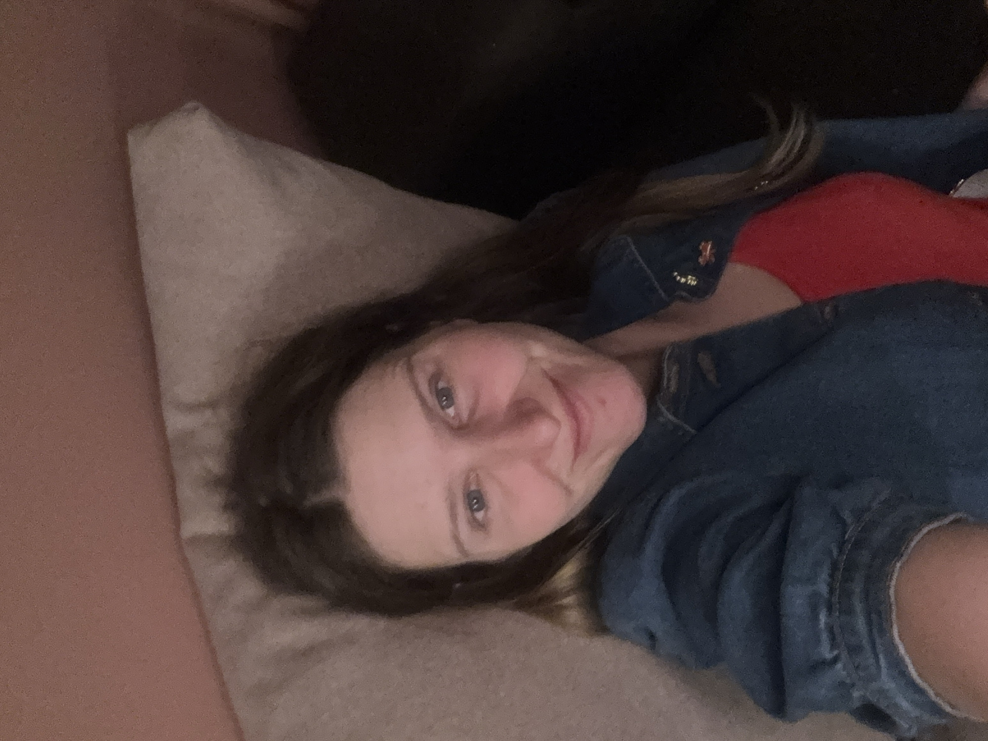 A woman lies on a light beige pillow against a pinkish wall. She is wearing a red top covered by a blue denim jacket. Part of a dark object is visible in the background. The location is Unknown, Unknown.