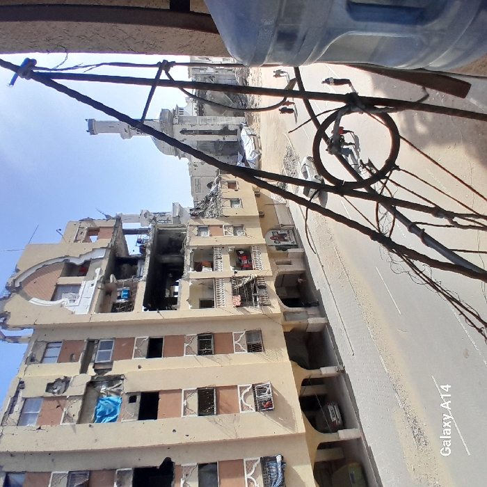 The image depicts a severely damaged urban street scene in Khan Yunis, Palestinian Territory, under a clear daylight sky. In the immediate foreground on the right, a partially visible translucent blue water cooler bottle is situated behind a dense tangle of rusty metal rods and wires, which obstructs part of the view.
To the left, a multi-story building, likely residential, exhibits extensive structural damage. Large portions of its tan-colored facade are blown out, revealing interior rooms and collapsed sections. Numerous impact marks or pockmarks are visible across the exterior walls. Blue fabric hangs from an upper-level window, and laundry is draped on a lower balcony.
The street, paved and appearing dusty with scattered debris, runs horizontally across the midground. A light-colored car is stationary or parked on the street. Further down, a greenish car is parked beneath an arched ground-level section of the damaged building. A small group of approximately three individuals stands near the light-colored car, interacting or observing. Another group of two people is walking further down the street towards the background.
In the background, a prominent mosque, identifiable by its distinctive minaret with a pointed dome, stands heavily damaged. The main structure of the mosque shows significant collapse and rubble, while the minaret itself, though scarred, remains largely upright. The overall scene indicates widespread destruction across the urban landscape.