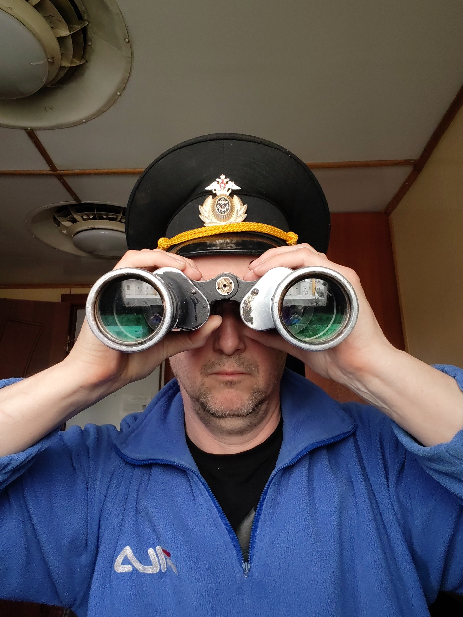 A man wearing a naval officer's hat and a blue fleece jacket is looking through a pair of binoculars. The hat is black with a gold trim and a white emblem. The jacket has a partial zipper and a white logo on the lower left. Reflected in the binoculars are images of a building, possibly located in Rybinsk, Russia, indicated by a window view and potentially recognizable architectural features if viewed closely. The background shows a ceiling with air conditioning units. The man is indoors.