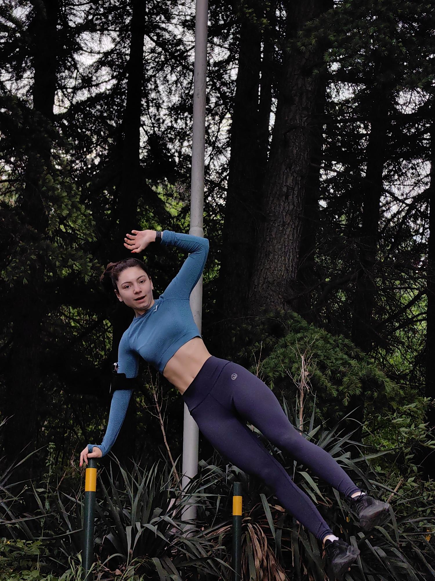 A woman in athletic attire is performing a side plank in a park-like setting. She is wearing a long-sleeved blue crop top and dark leggings, with an armband on her left arm. Her body is angled, supported by her left hand on a green and yellow post, and her feet are resting on another post. The background consists of dense trees and greenery. The location is Varna, Bulgaria, as evidenced by the specific vegetation, architecture, and terrain typical of parks within the city.