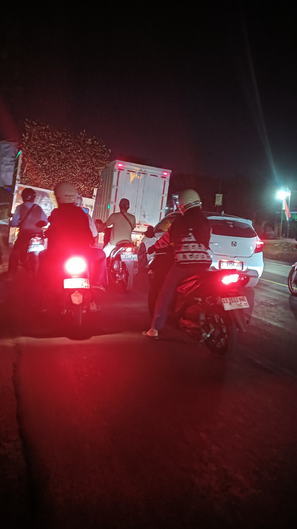 The image depicts a nighttime traffic scene in Ngrampal, Indonesia, possibly on Jalan Raya Ngrampal. Several motorcycles, a white car, and a large white truck are visible, suggesting a traffic jam or slow-moving traffic. The lighting conditions are poor, with the primary illumination coming from the vehicles' headlights and taillights, casting red and white glows across the asphalt road.
In the foreground, three motorcycles are prominent. The closest one on the left has its bright red taillight illuminated. A person wearing a light-colored shirt and a helmet is seated on this motorcycle, facing away from the camera. To the right of this, another motorcycle has its taillight on, and a person in a dark jacket and helmet is riding it, also facing away. A third motorcycle on the far right has its taillight visible, and a person is seated on it.
In the middle ground, a white car with a license plate ending in "QV" is positioned behind the motorcycles. Further back, a large white truck is also present, its rear lights visible. Another motorcycle is seen between the white car and the truck, with a person wearing a light-colored shirt and a cross-body bag.
The environment appears to be a road with some foliage or trees visible on the left side of the image, illuminated by the truck's lights. The overall impression is one of congestion and slow movement on a dark road, typical of a busy intersection or a stretch of road experiencing heavy traffic.