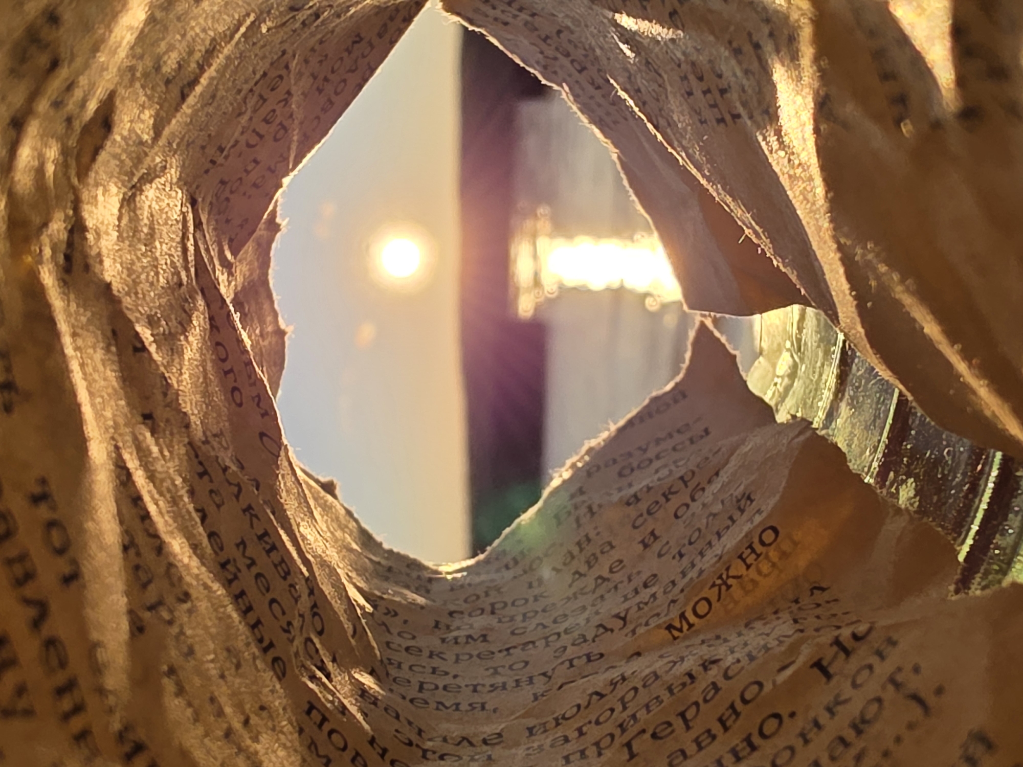 The image shows a view through a torn piece of paper, likely from a bag or packaging material with Cyrillic text printed on it. The tear forms a roughly diamond-shaped opening through which the sun shines brightly. In the background, indistinct shapes and reflections suggest an urban setting, possibly in the city of Chaikovsky, Russia, as indicated by the Cyrillic writing. The paper edges are crumpled and textured, and the light creates a warm, golden tone throughout the scene. The focus is on the light and the background, with the paper acting as a frame.